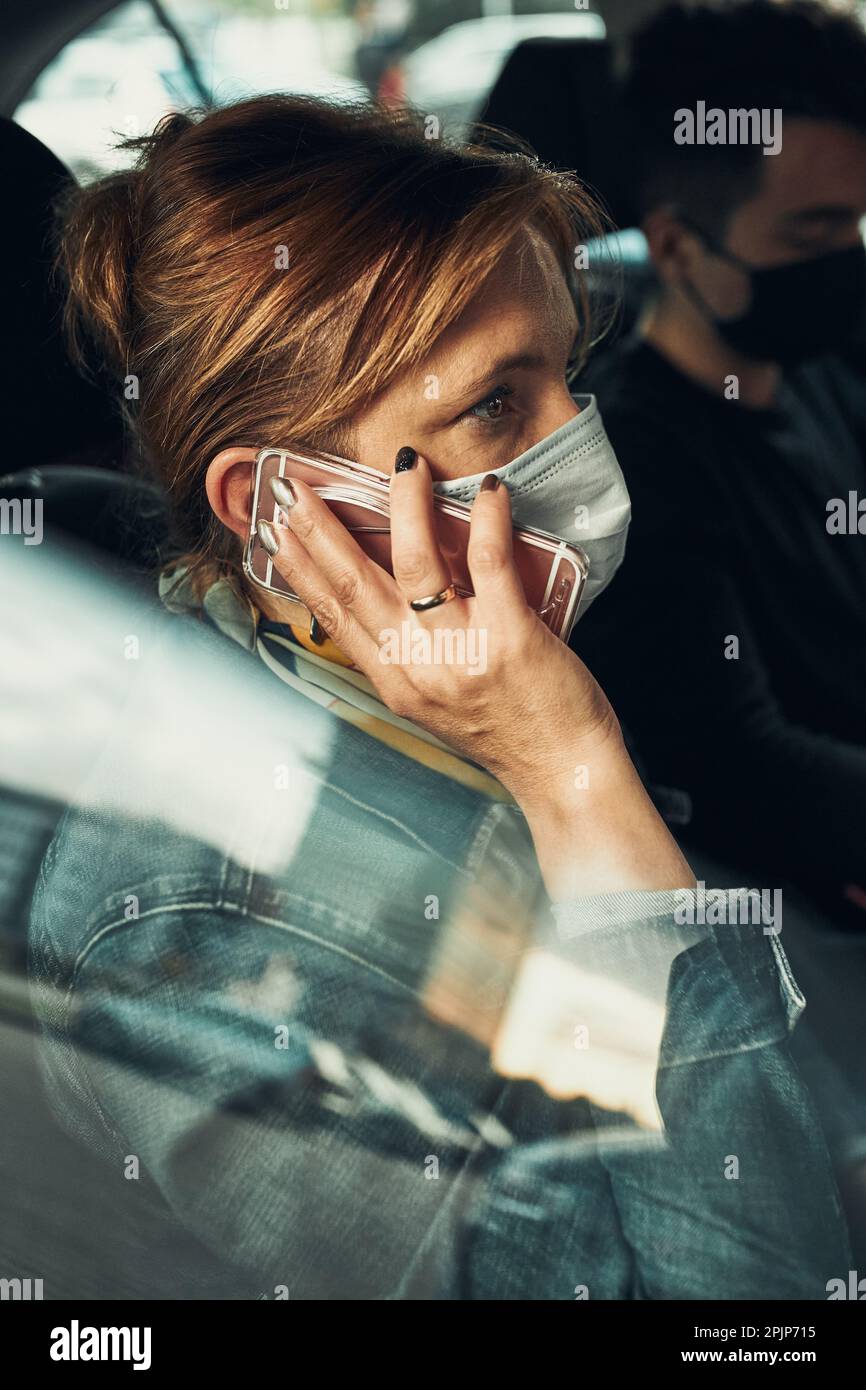 Donna che parla al telefono utilizzando uno smartphone seduto in un'auto che indossa la maschera facciale per evitare l'infezione da virus e per prevenire la diffusione della malattia in tempo o Foto Stock