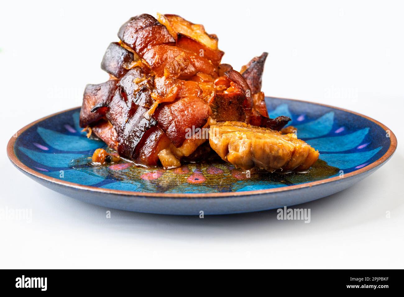 Pancia di maiale al forno e aglio su piatto blu decorativo, spicchio su sfondo bianco. Foto Stock
