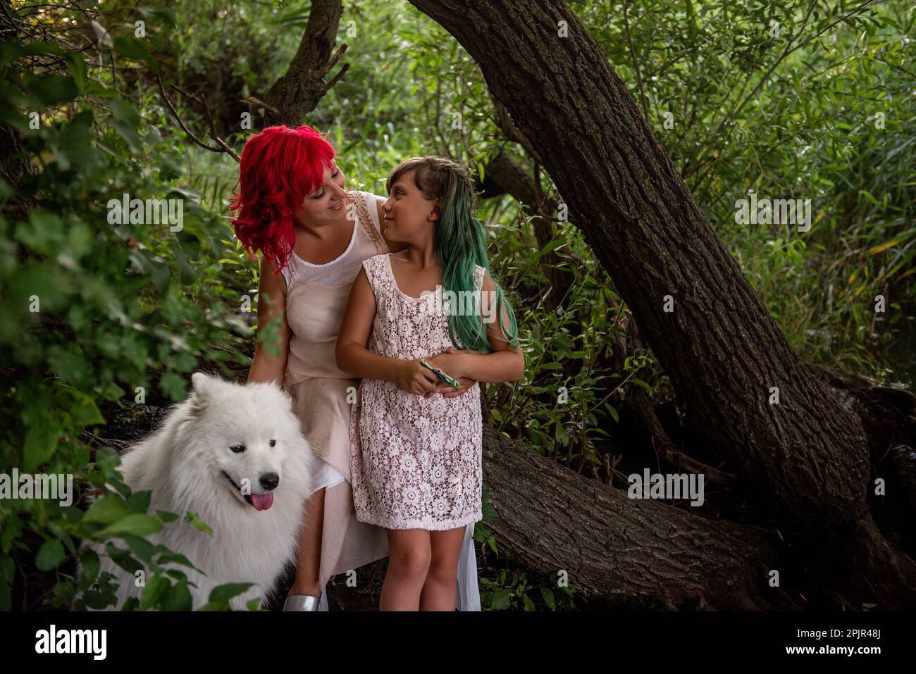 Nella Foresta Verde, da piccolo ruscello diversità, madre con capelli rosa abbraccia figlia. Il bianco soffice samoyed custodisce la famiglia. In viaggio con animali domestici Foto Stock