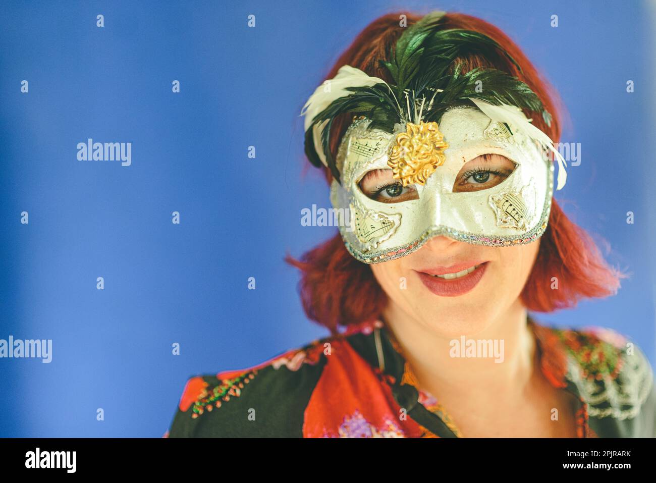 Ritratto di una donna adulta con testa rossa caucasica che indossa una maschera teatrale veneziana, studio spara terra blu con messa a fuoco selettiva Foto Stock
