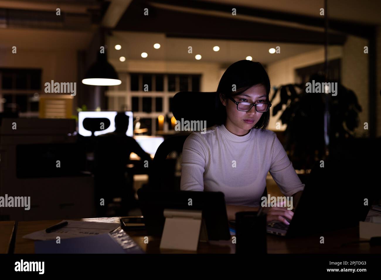 Donna d'affari asiatica che indossa occhiali, seduto alla scrivania, con un computer portatile che lavora tardi in ufficio Foto Stock