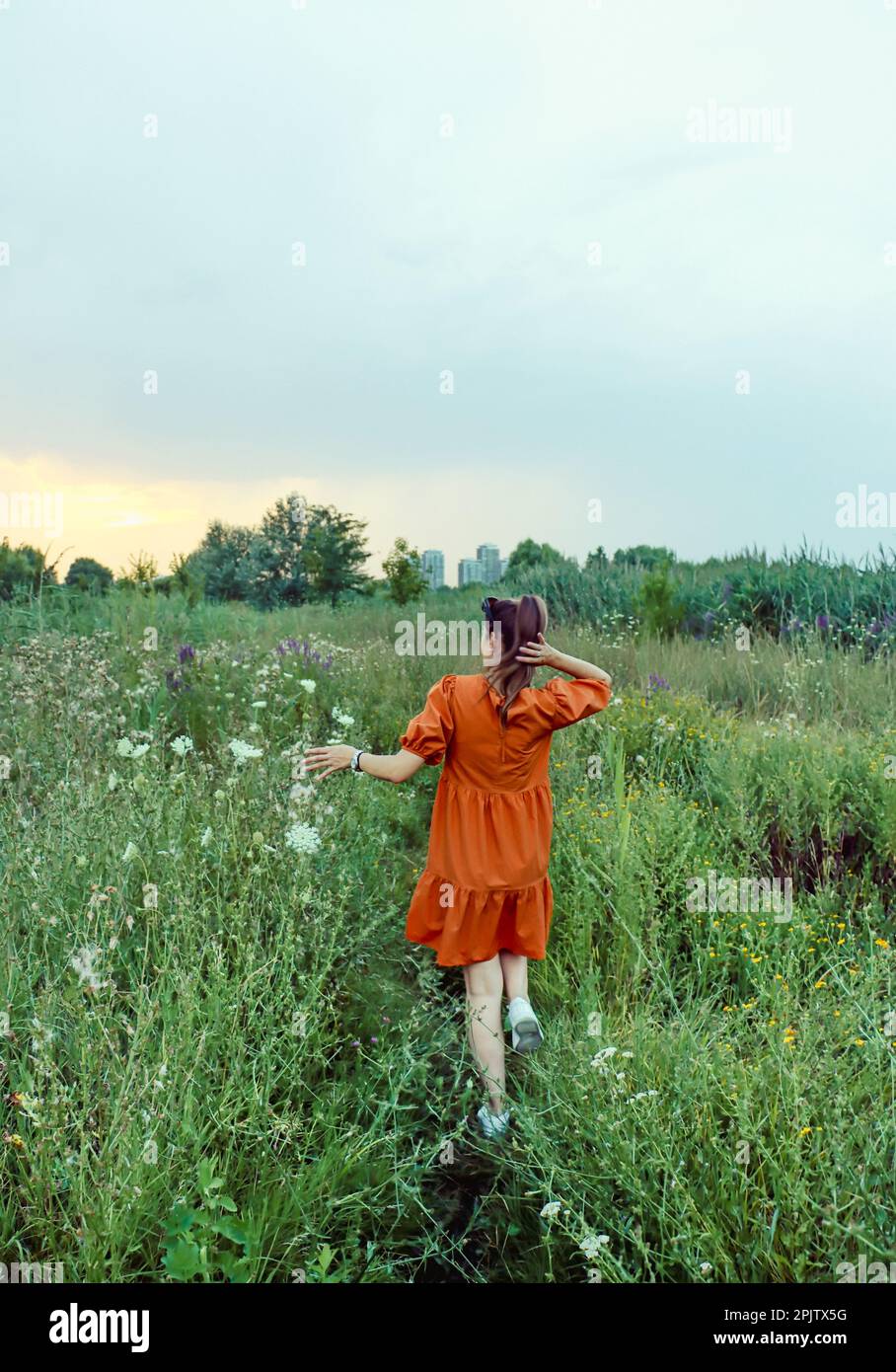 Ragazza in un abito arancione che viaggia da sola attraverso il campo dei fiori primaverili, ammirando i colori del tramonto Foto Stock