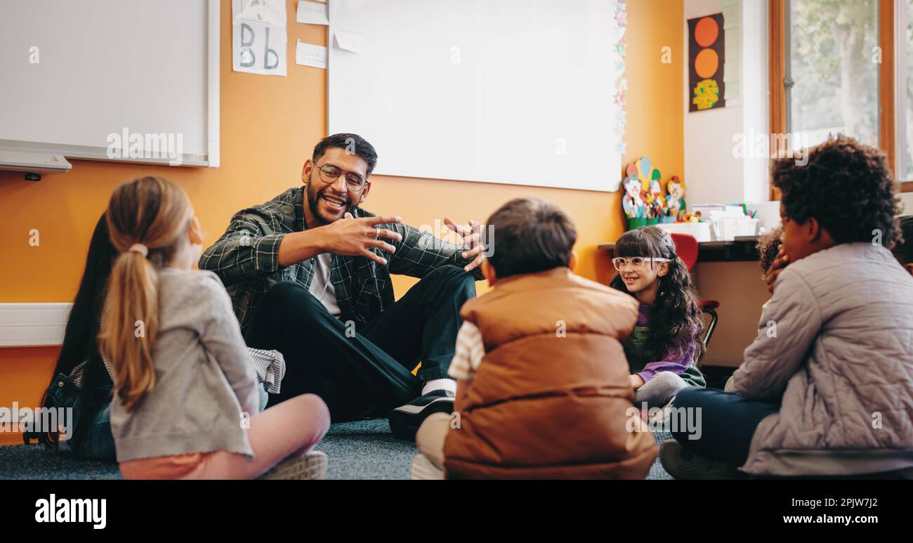 Educatore maschio che parla con i suoi studenti in una classe. Uomo che insegna ai bambini delle scuole elementari, è seduto sul pavimento con loro e dà una verba Foto Stock