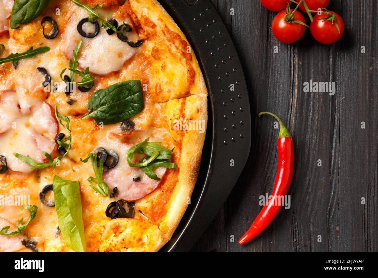 Pizza con jerky, olive, formaggio e verdure su un piatto nero rotondo e verdure fresche su uno sfondo di legno scuro. Pizza fatta in casa con carne ed erbe Foto Stock