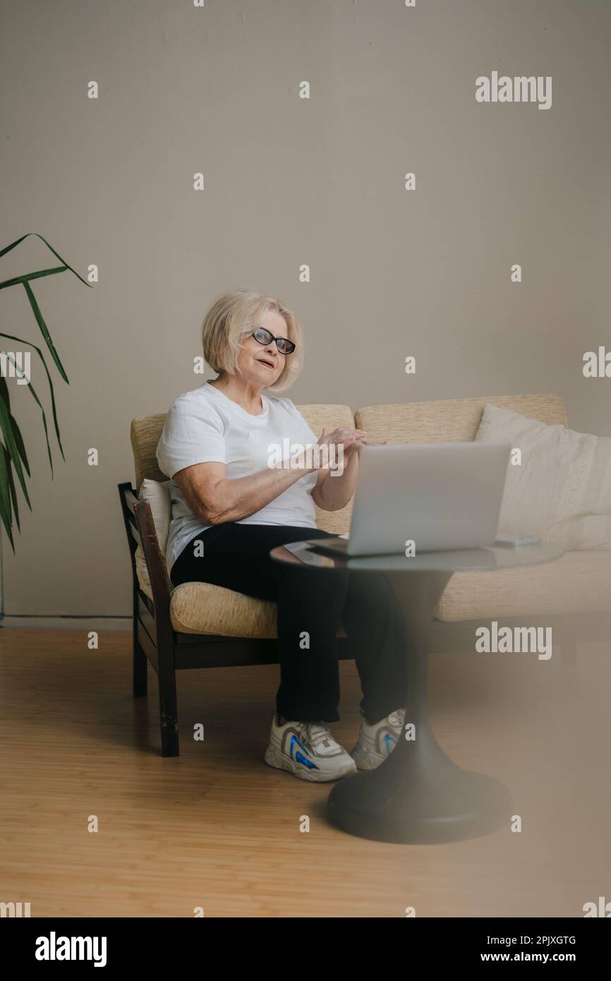 Donna anziana che applica crema cosmetica sulle mani mentre lavora lontano da casa utilizzando un dispositivo portatile, sorridendo allegro. Concetto di bellezza. Persone Foto Stock