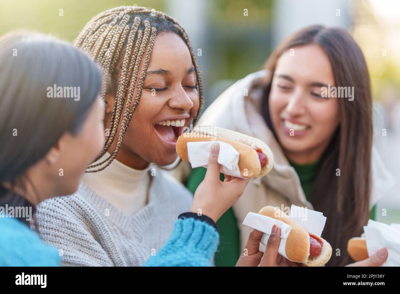 Tre amici sorridenti mangiano hot dog in un parco Foto Stock