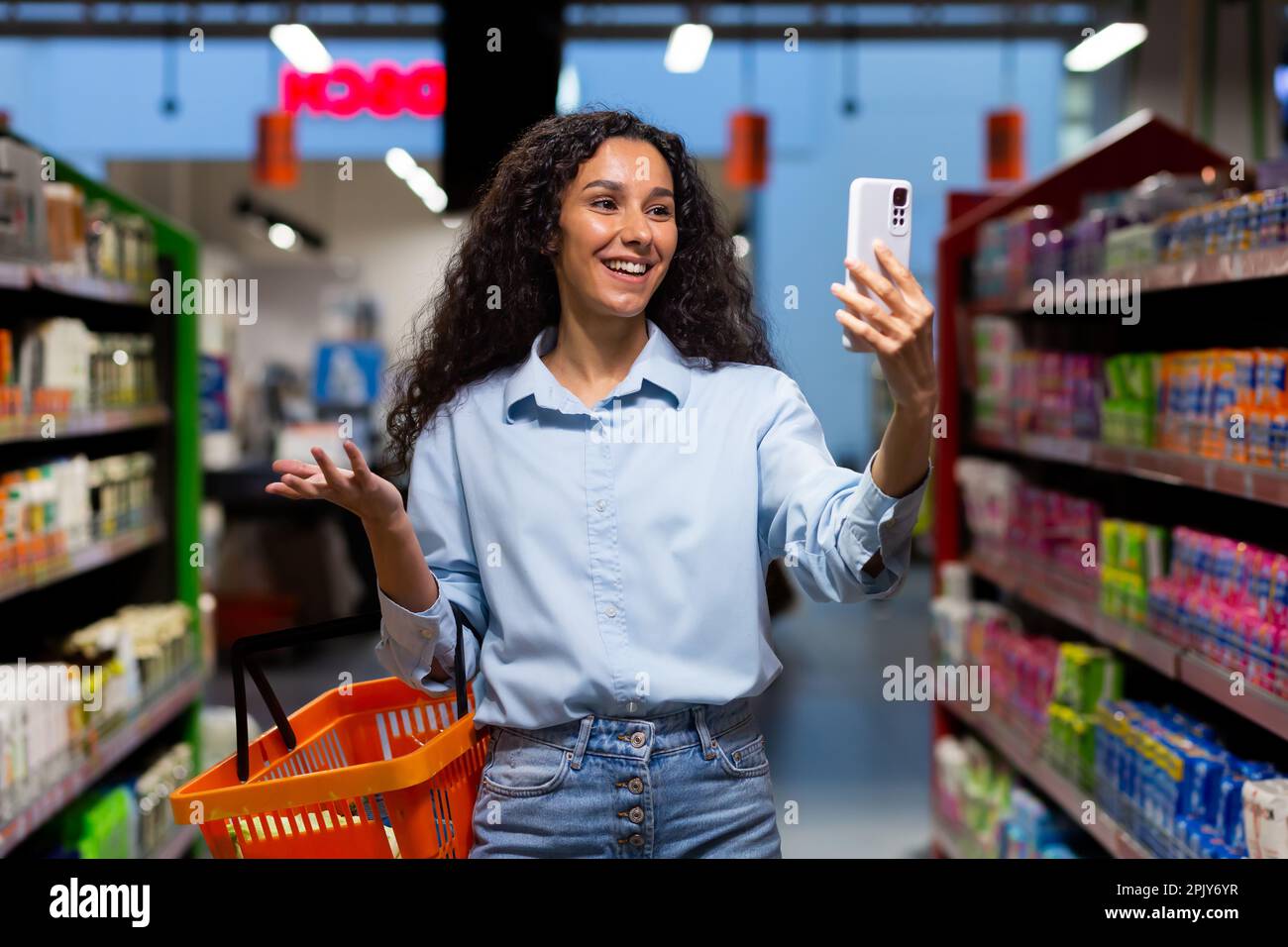Una giovane donna latino-americana, blogger si trova in un supermercato con un cesto tra gli scaffali con le merci e parla di una videochiamata dal telefono, consulta, chiede cosa comprare, sorride. Foto Stock