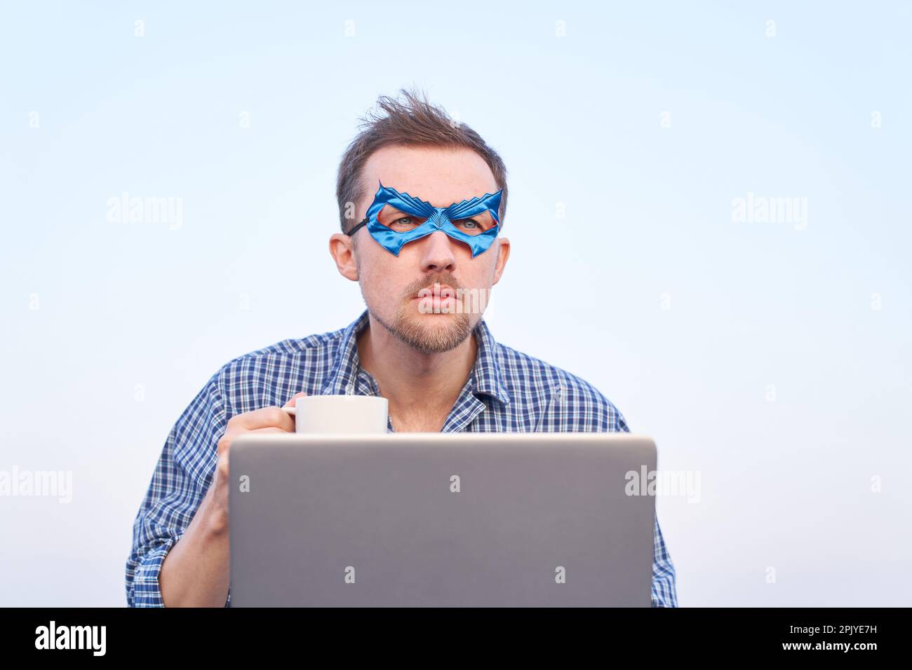 Uomo allegro in camicia da notte e maschera viso blu con laptop all'aperto. Supporta il supereroe che utilizza un computer portatile con una tazza di tè o caffè caldo. Immagini di alta qualità Foto Stock