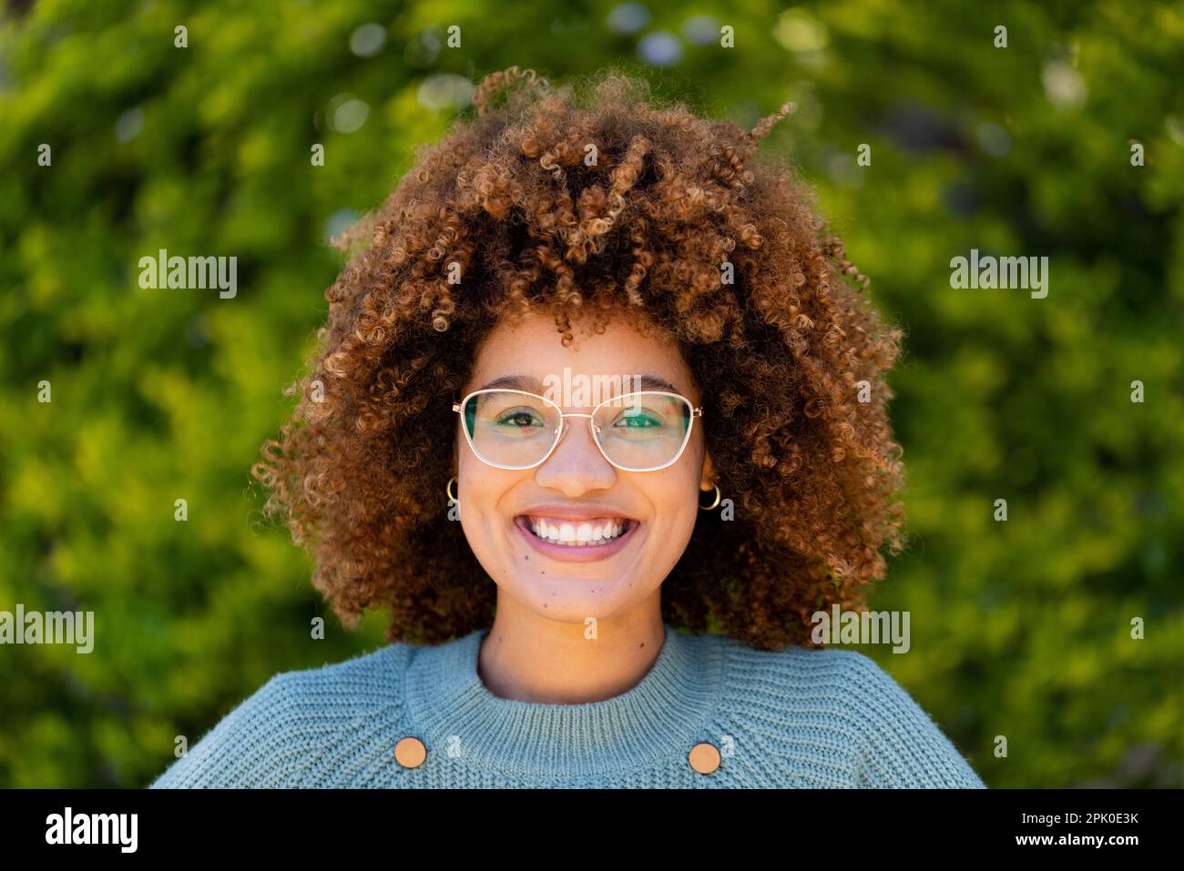 Primo piano ritratto di bella giovane donna biraciale con capelli afro sorridente contro le piante in cortile Foto Stock