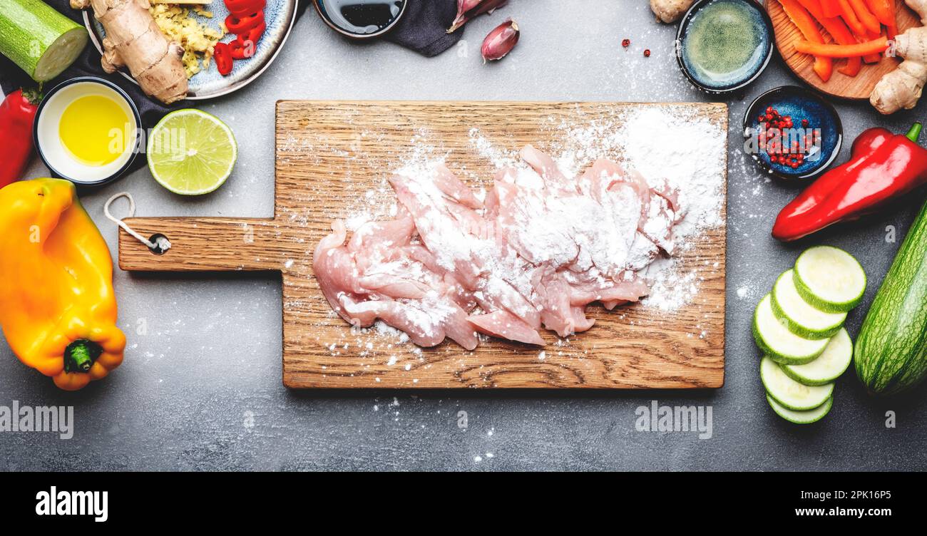Cibo sfondo. Filetto di pollo crudo a fette cosparso di amido sul tagliere, verdure, zenzero, lime, peperoncino, aglio, salsa di soia e riso Foto Stock