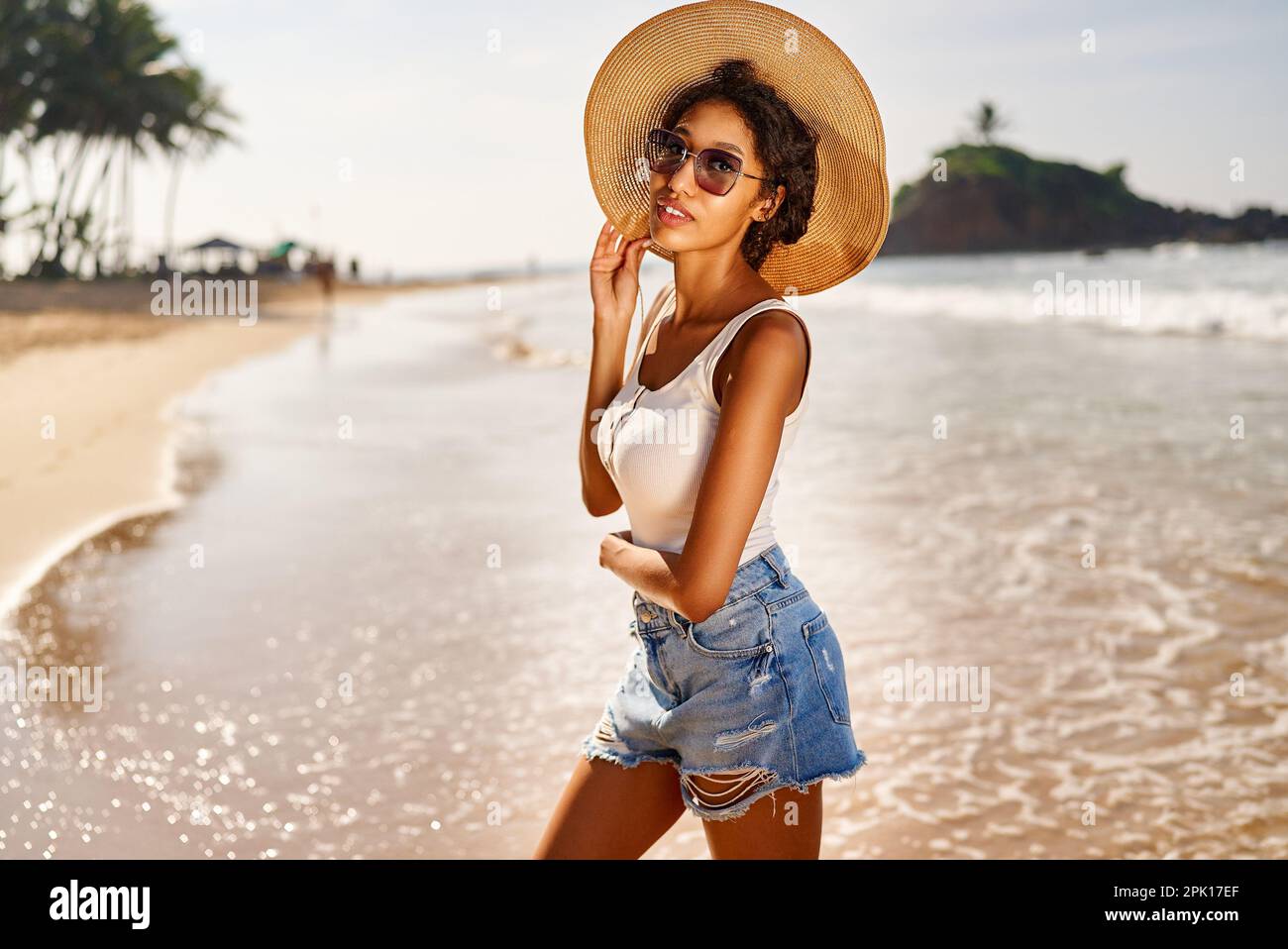 Giovane donna africana modello in cappello di paglia e occhiali da sole in posa presso il resort dal mare all'alba. Donna nera contro l'isola rocciosa e l'oceano Foto Stock