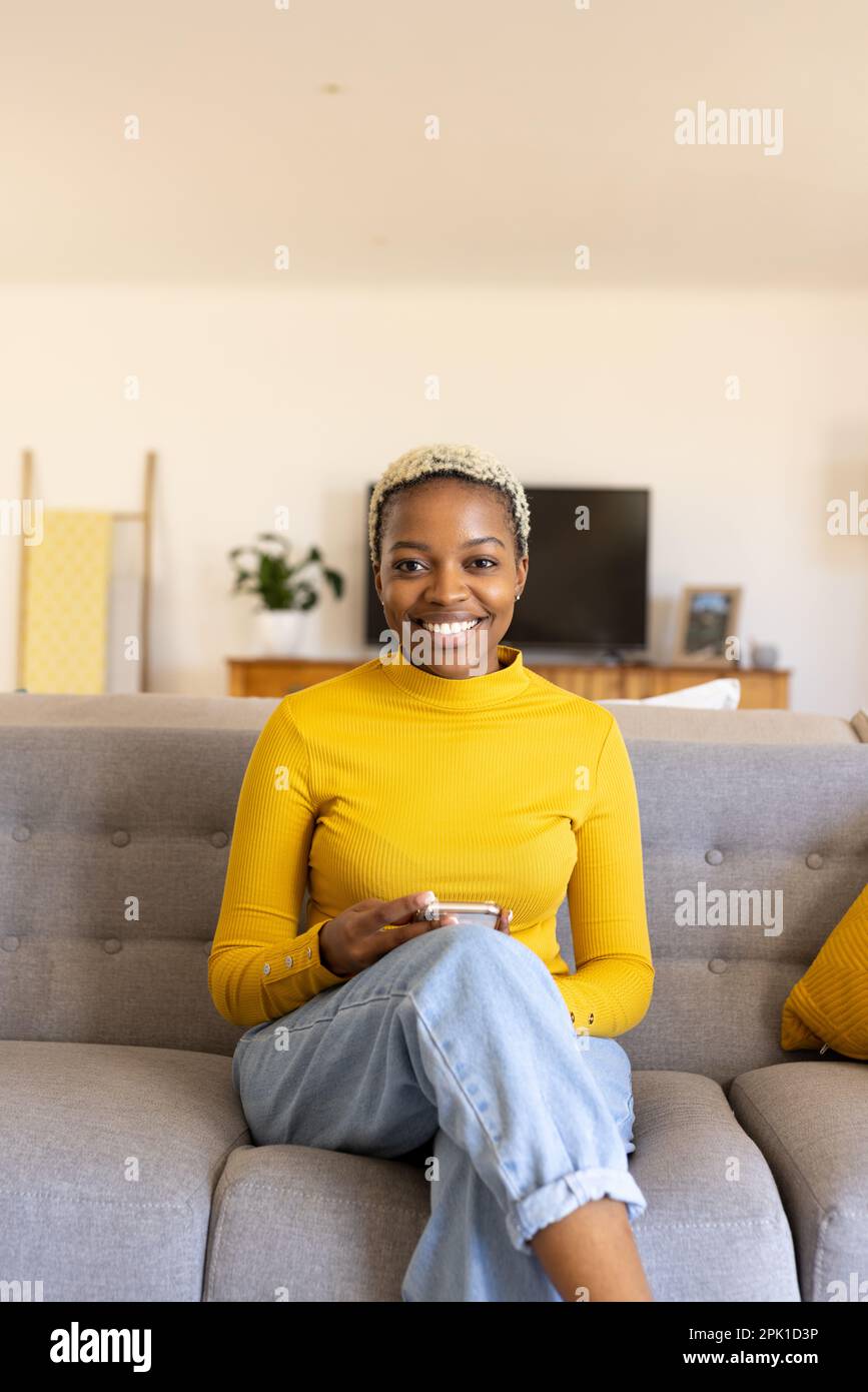 Ritratto di giovane donna afroamericana con capelli corti utilizzando smartphone mentre si siede sul divano Foto Stock