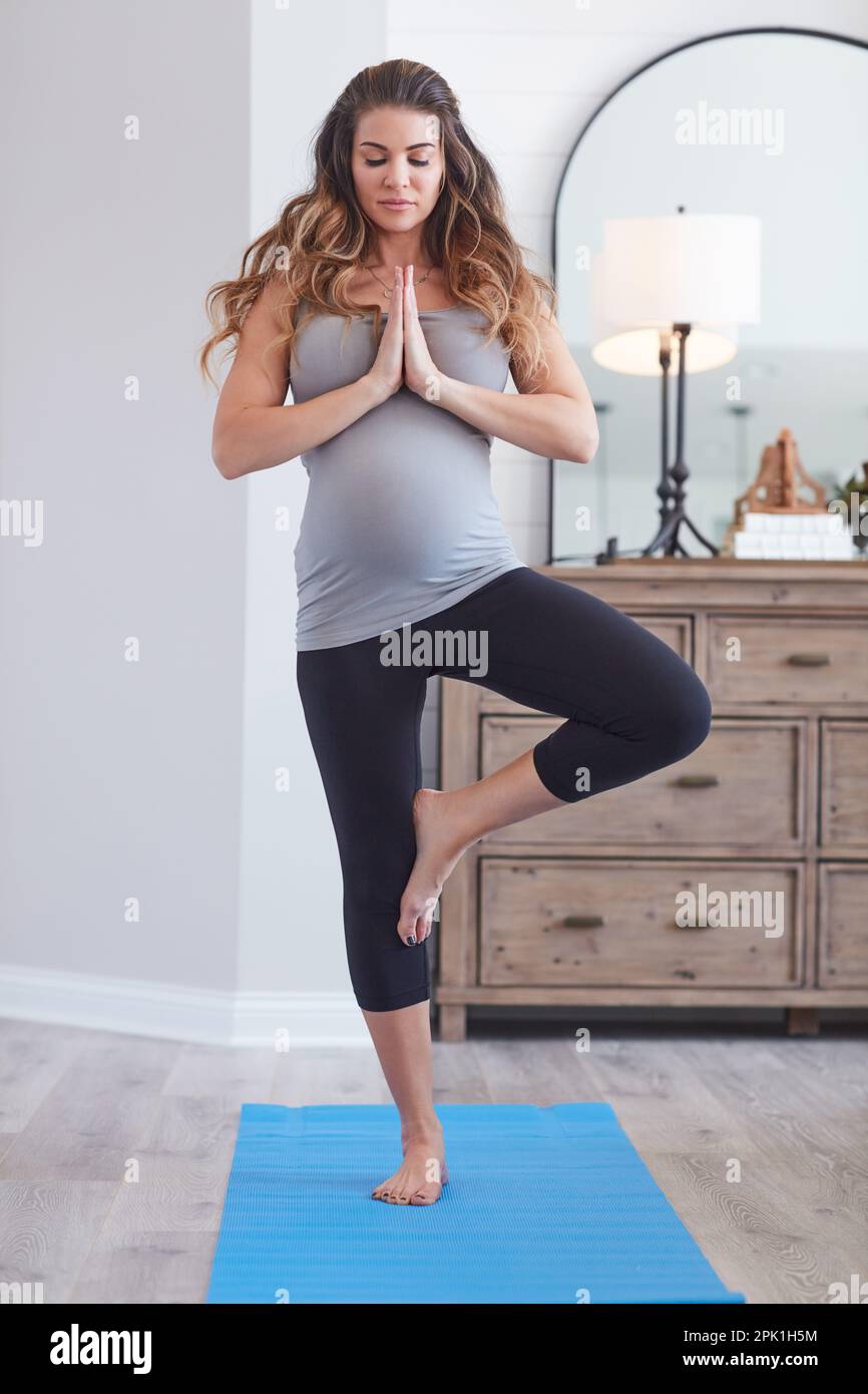 Preparandosi per il più grande atto di equilibrio della sua vita. una giovane donna incinta che pratica la posa dell'albero durante una procedura di yoga a casa. Foto Stock