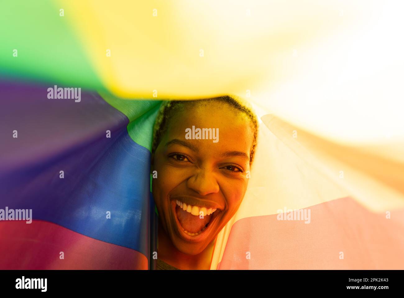 Primo piano di allegra giovane donna afroamericana con capelli corti che urlano sotto la bandiera arcobaleno Foto Stock