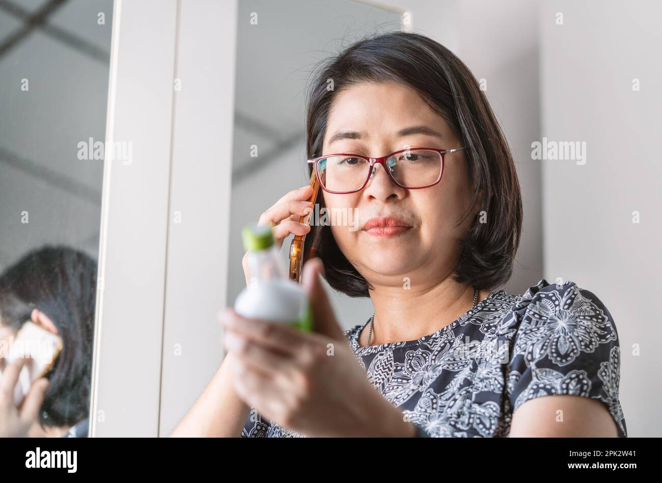 La donna asiatica sta ascoltando un medico consiglia l'uso medico dal suo smartphone, la sua mano sta tenendo un flacone di medicinale liquido, gli occhi che guardano il flacone. Foto Stock