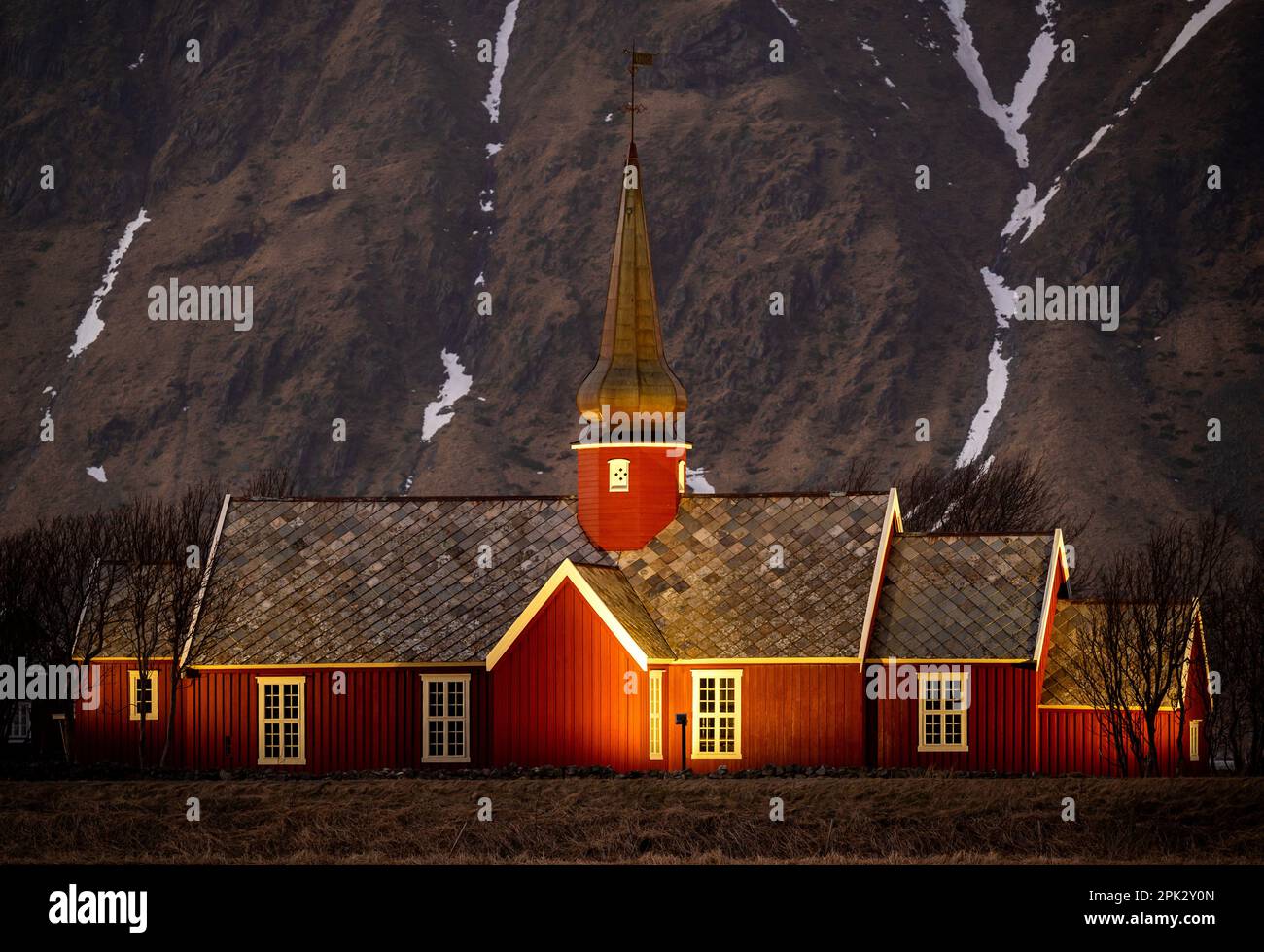 Chiesa in legno di colore rosso da 1780 anno, illuminata di sera con alta montagna rocciosa con neve. Flakstad, Lofoten, Norvegia. Foto Stock