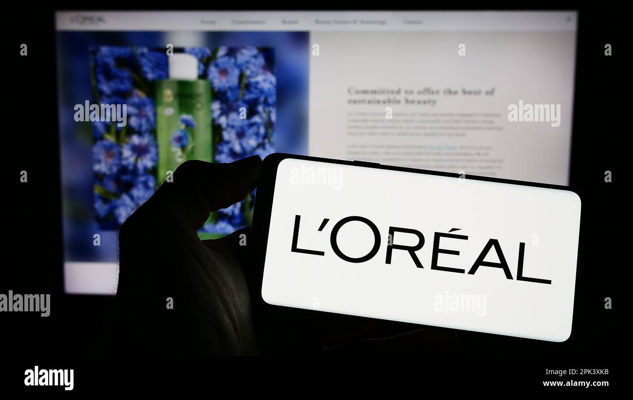Persona che tiene il cellulare con il logo della società francese di cura personale l'Oreal SA sullo schermo di fronte alla pagina web aziendale. Messa a fuoco sul display del telefono. Foto Stock