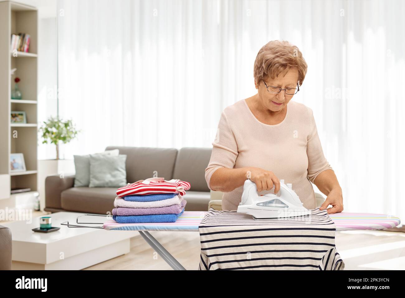 Donna anziana che stira i vestiti a casa in un soggiorno Foto Stock