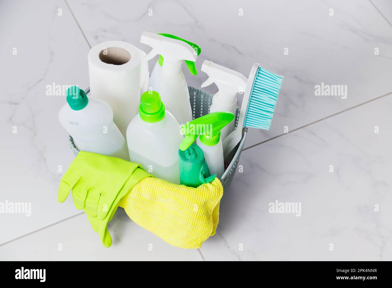 Cestino con prodotti per la pulizia su pavimento in ceramica bianca in cucina. Diversi flaconi di detergente, guanti di gomma, dispenser spray, tovaglioli e spazzola per Foto Stock