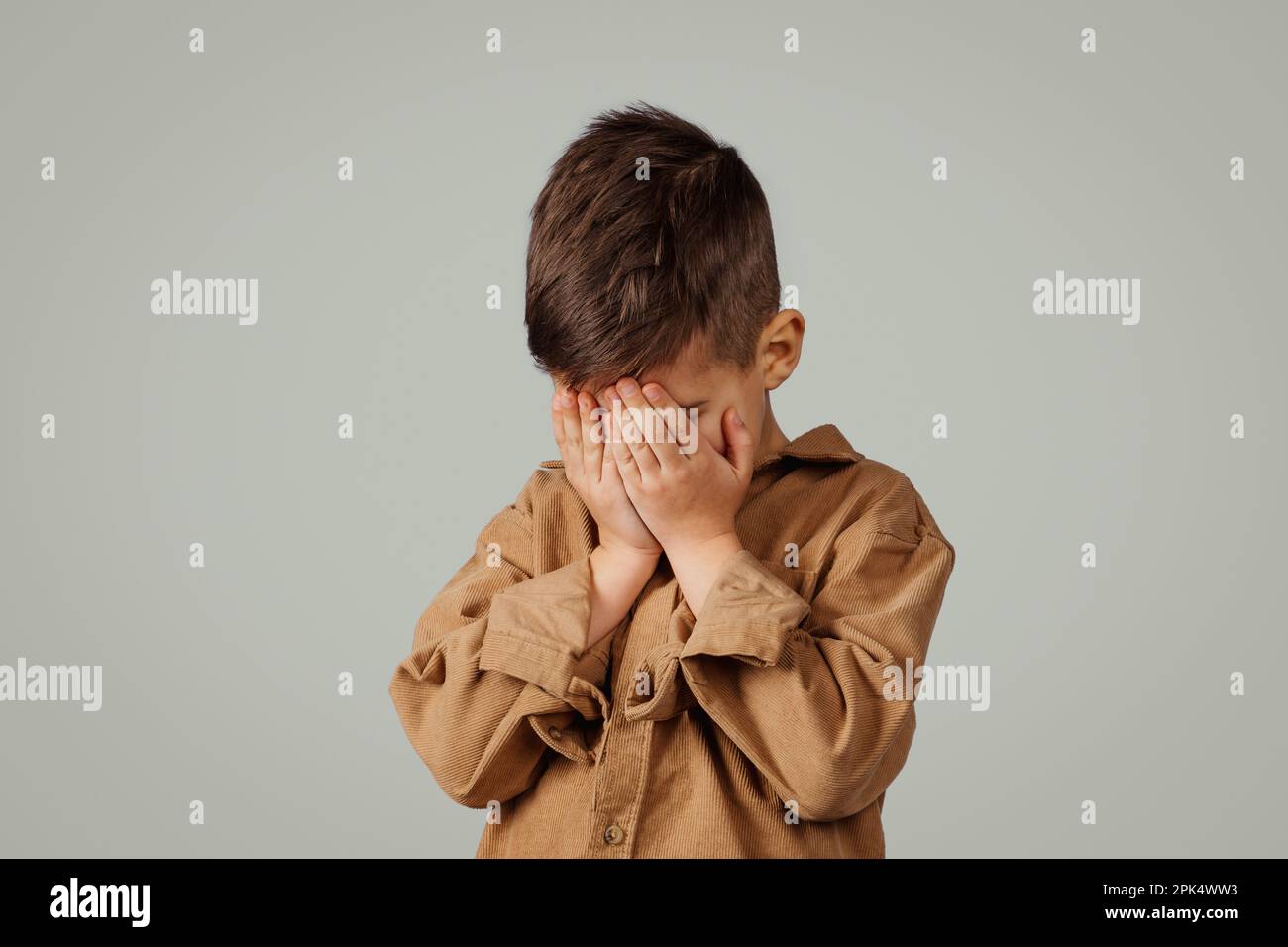 Triste caucasico bambino di 6 anni che copre il viso con le mani, piangendo isolato su sfondo grigio studio Foto Stock