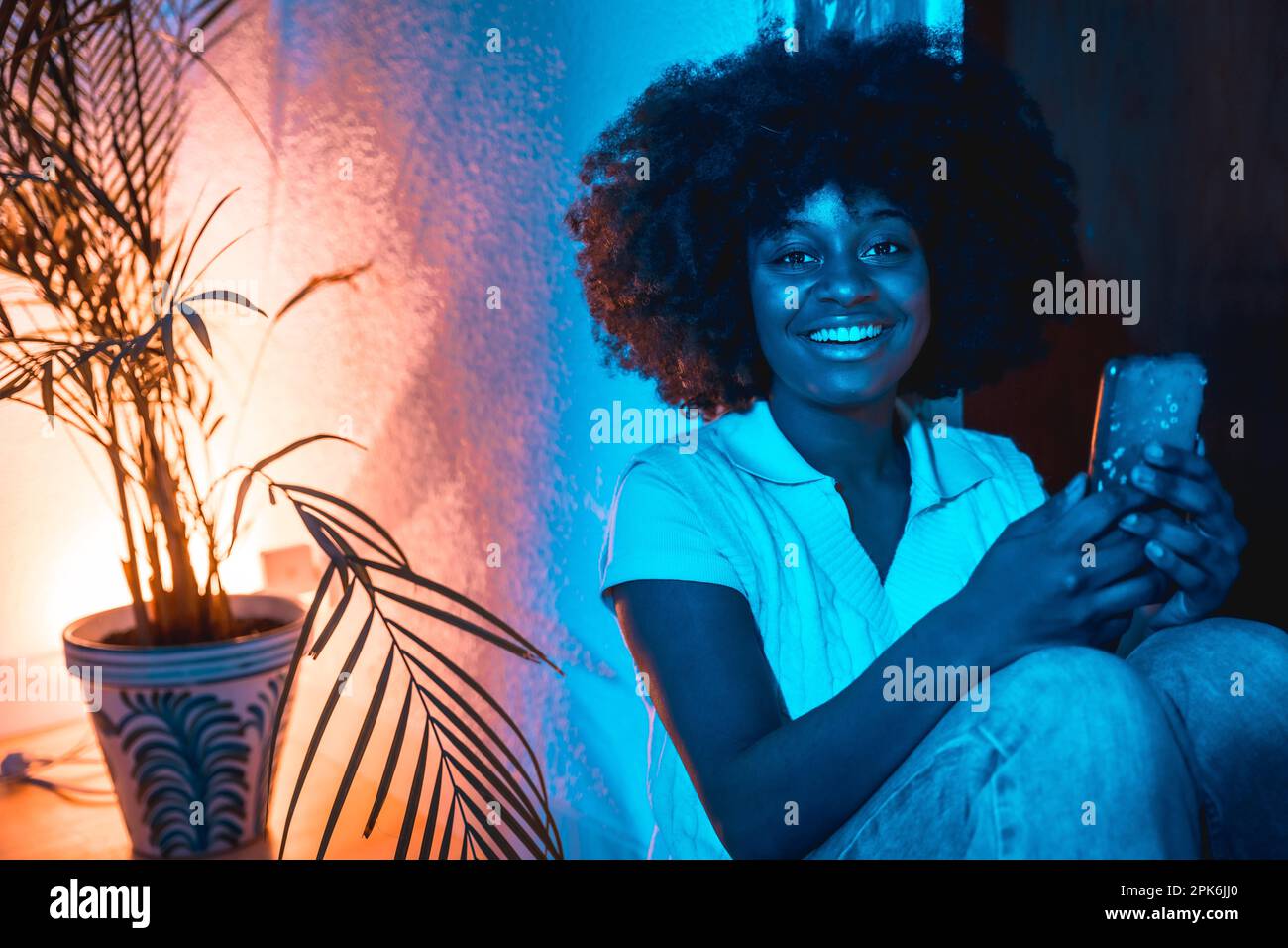 Donna afroamericana con capelli afro seduto a casa sul pavimento guardando il telefono di notte con una luce blu Foto Stock