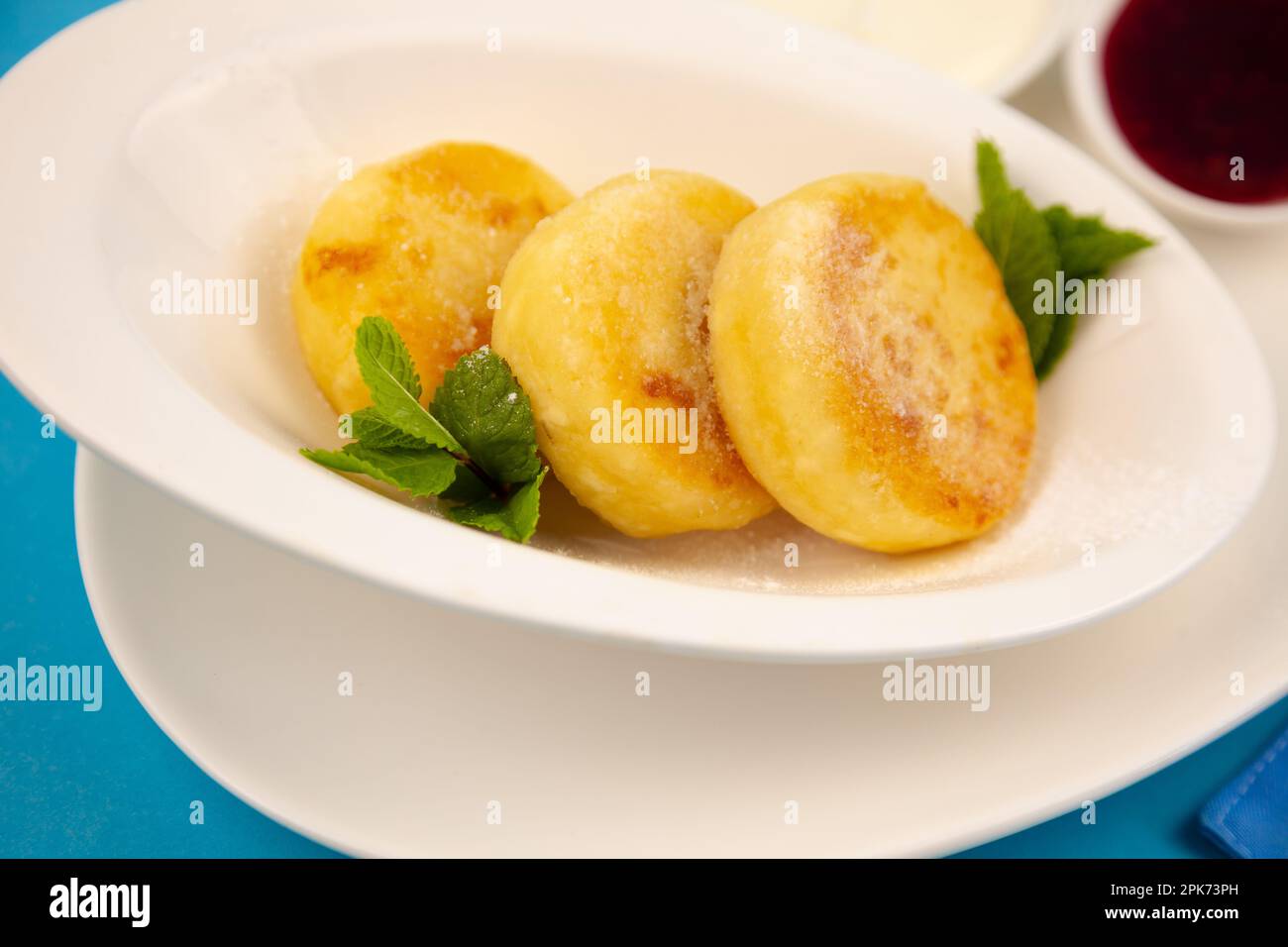 Frittelle di formaggio ricotta rotonda in un piatto bianco su sfondo blu. Cibo prima colazione. Cibo sano. Salsa di panna acida e menta. Primo piano vista dall'alto. Pasto dessert. Frittelle di ricotta al formaggio ricotta. Foto Stock