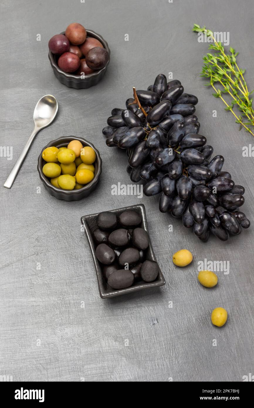 Olive in ciotole. Mazzo di uve nere, rametto di timo e cucchiaio sul tavolo. Giacitura piatta. Sfondo grigio Foto Stock