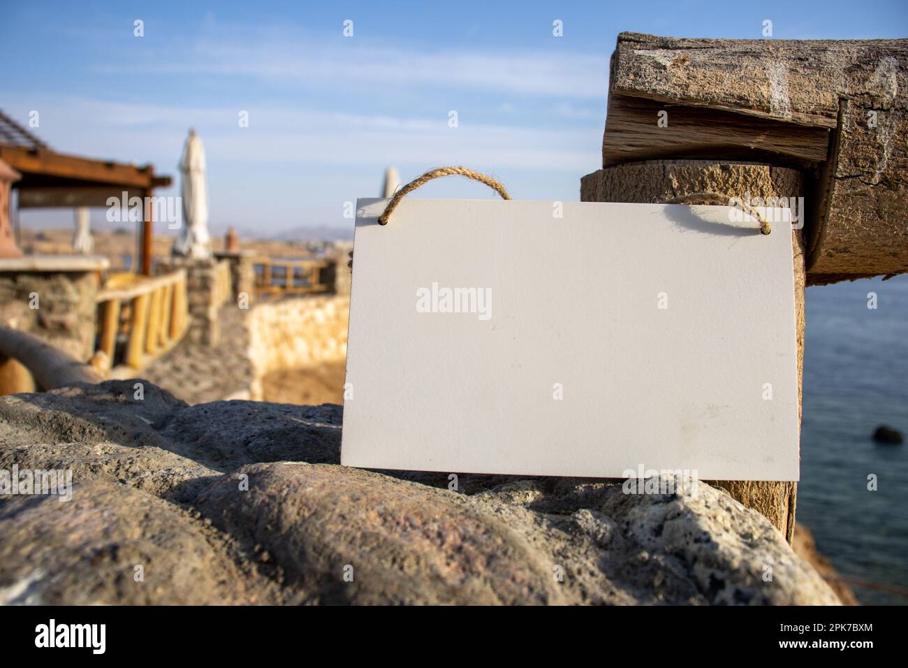 parete ruvida e recinzione con il mare sullo sfondo e cartello bianco sospeso con spazio libero vuoto per template o area di copia vuota Foto Stock