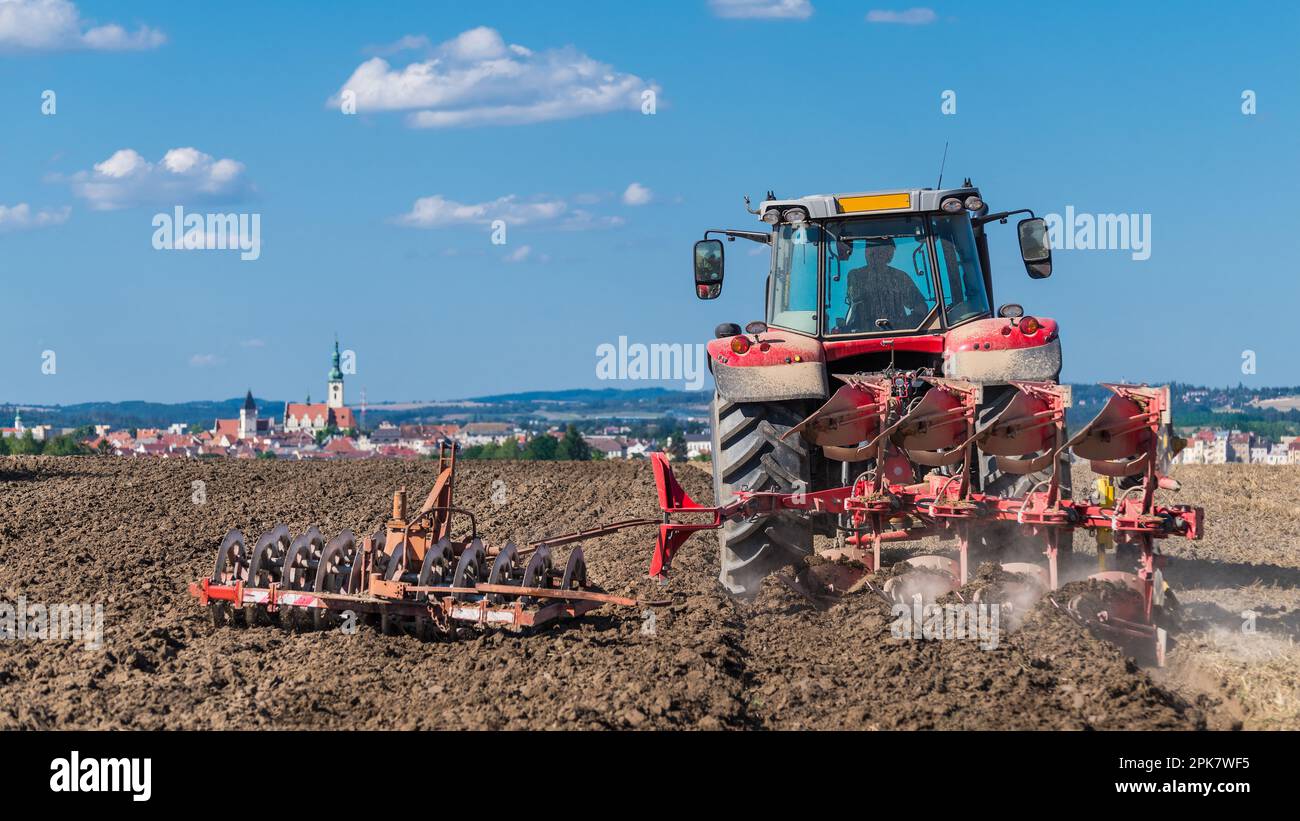 Trattore rosso con aratro e erpice a disco in terreno arabile marrone sotto il cielo blu estivo. Veicolo agricolo su campo polveroso e skyline della città ceca hussites Tabor. Foto Stock
