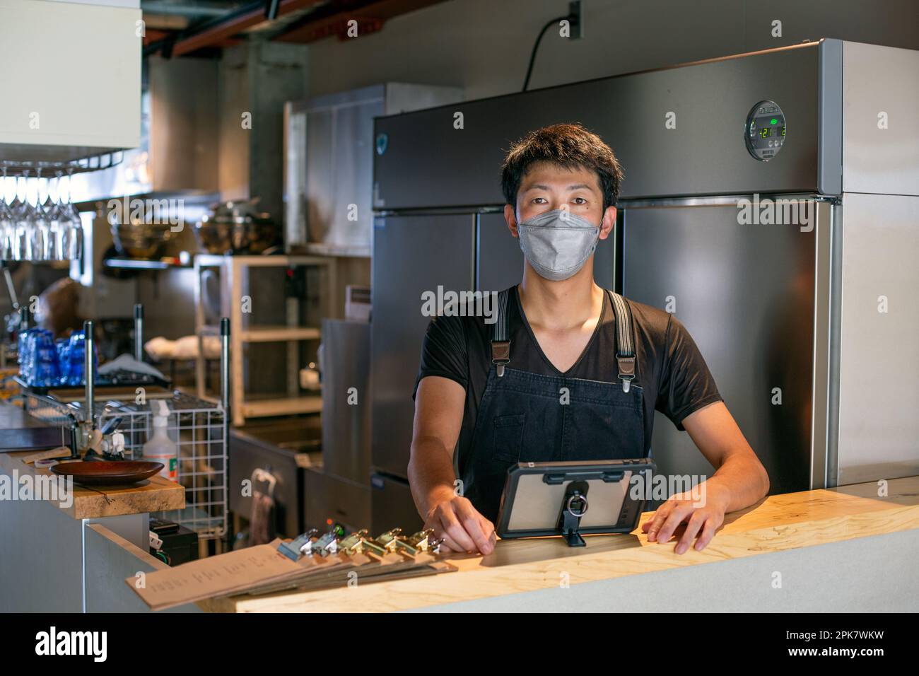 Un uomo che indossa una maschera al bancone di una cucina da ristorante, utilizzando un tablet digitale, il proprietario o il manager. Foto Stock
