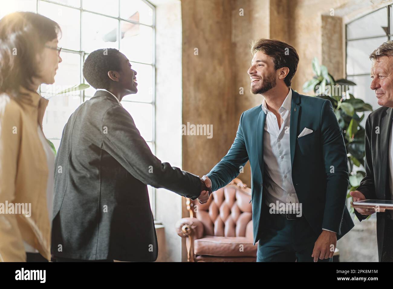 Giovane uomo caucasico e donna afroamericana con capelli rasati sorridendo e scuotendo le mani in un ufficio moderno, con i colleghi a guardare. Luce naturale Foto Stock