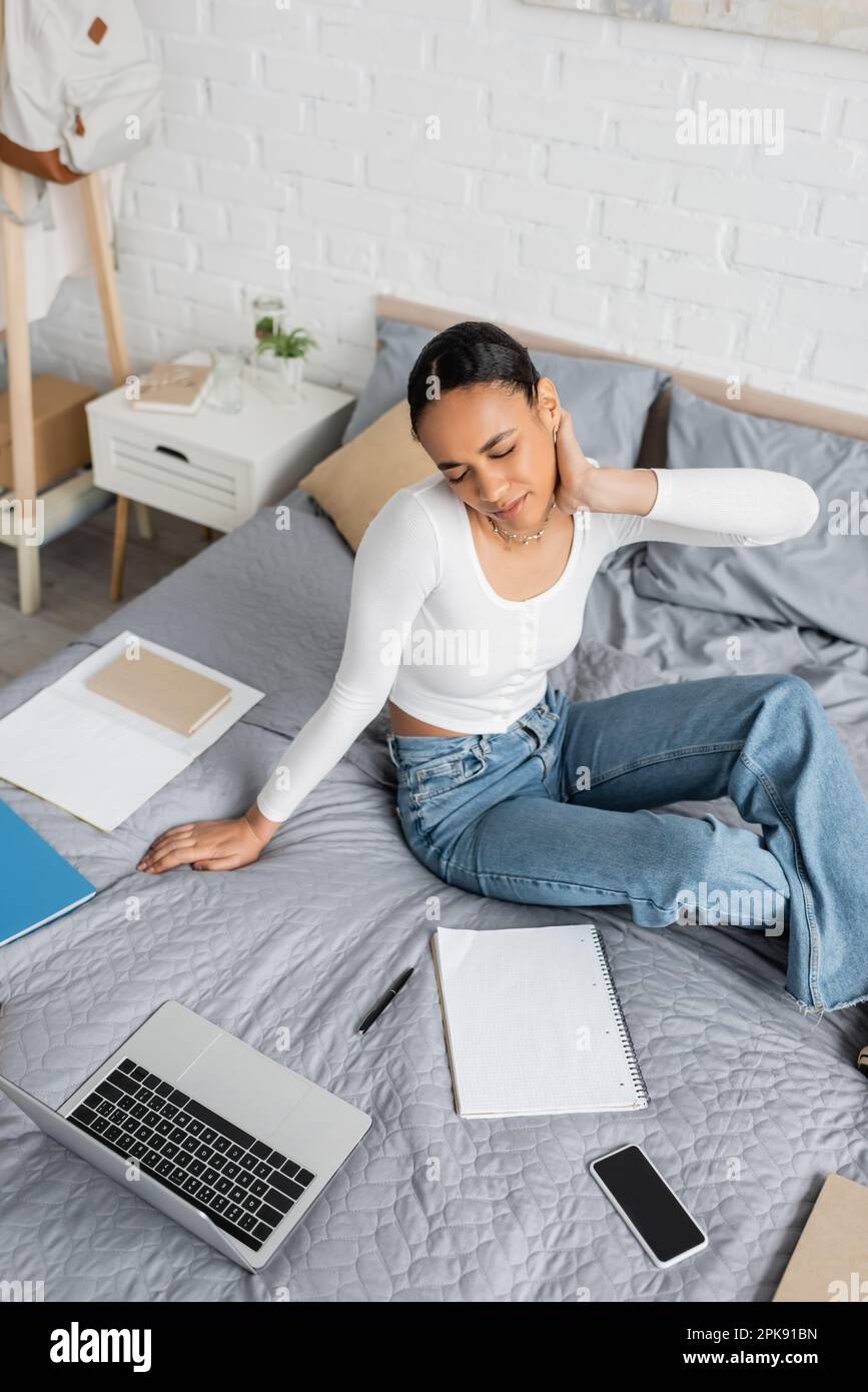 Studente africano americano stanco seduto vicino a notebook e notebook a letto, immagine stock Foto Stock