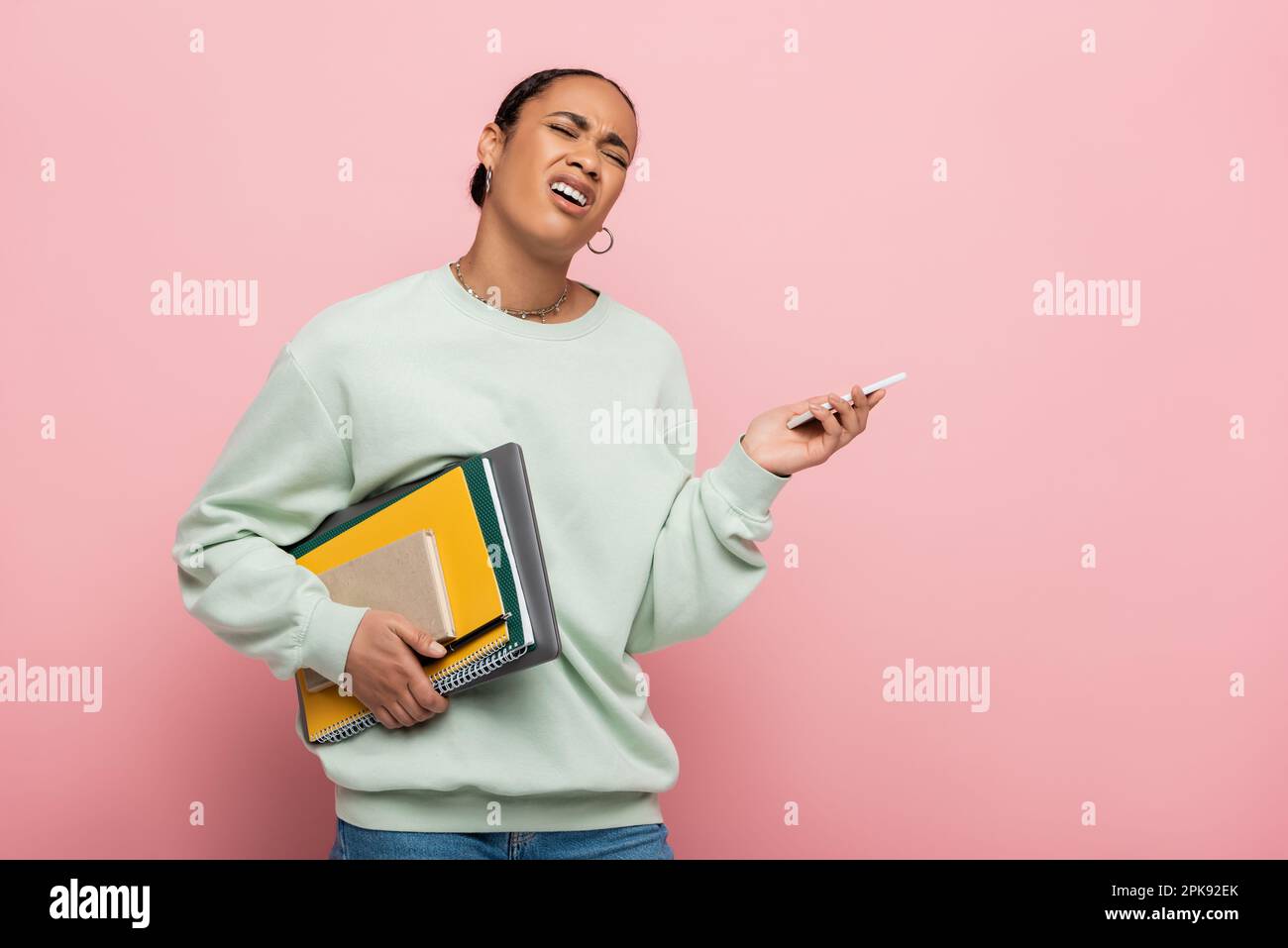 allievo afroamericano dispiaciuto in felpa che tiene i gadget e le forniture di studio mentre gemendo isolato su rosa, immagine di scorta Foto Stock