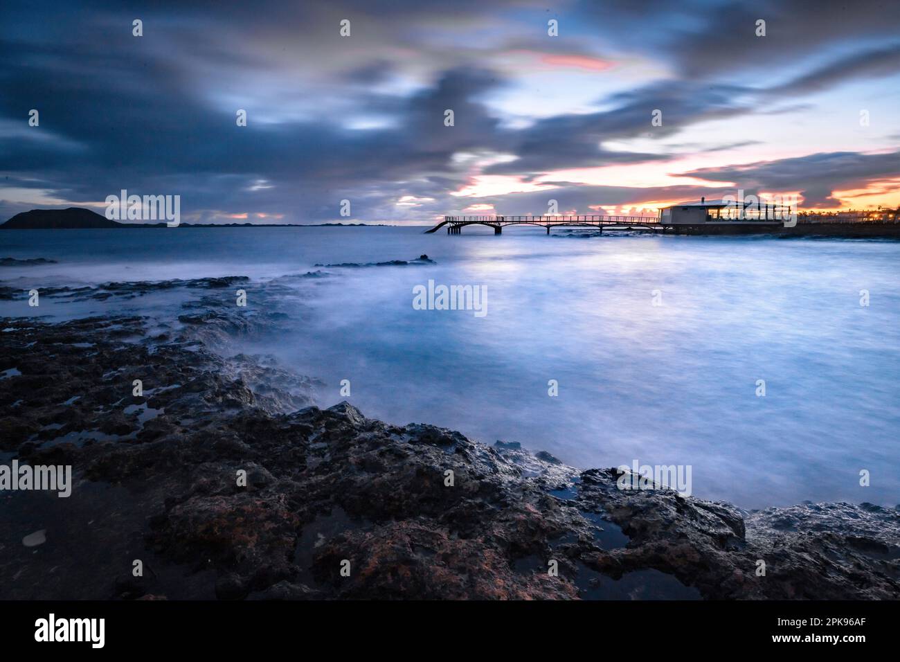 Alba sulla spiaggia, rocce laviche e spiaggia sabbiosa in lunga esposizione. Posto romantico con molo al mattino. Playa de la Barreta, Parco Nazionale di Corralejo, Isole Canarie, Spagna Foto Stock