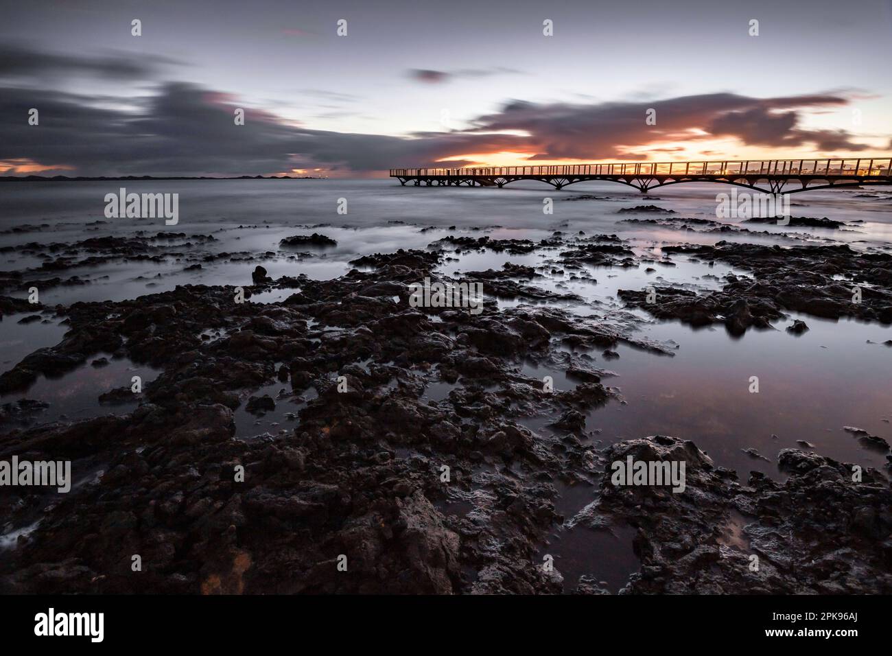 Alba sulla spiaggia, rocce laviche e spiaggia sabbiosa in lunga esposizione. Posto romantico con molo al mattino. Playa de la Barreta, Parco Nazionale di Corralejo, Isole Canarie, Spagna Foto Stock