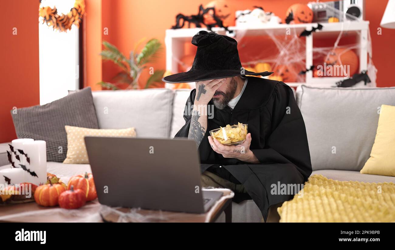 Giovane uomo calvo che indossa costume da mago mangiare patatine patate guardando il film a casa Foto Stock