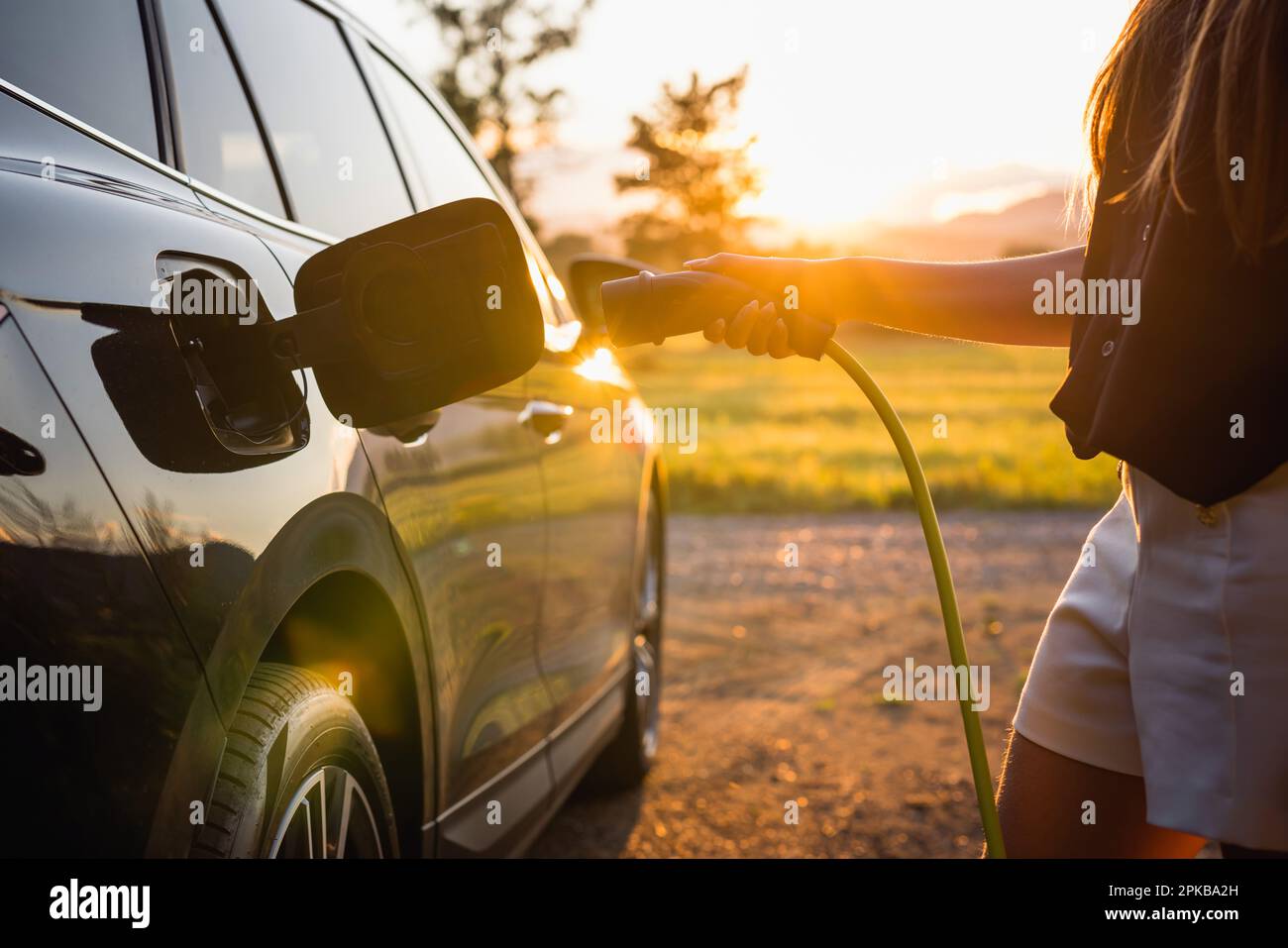 Mano femmina che mette un caricatore di cavo in un'automobile elettrica illuminata dai raggi del sole che rompono attraverso i rami dell'albero frondoso, primo piano Foto Stock