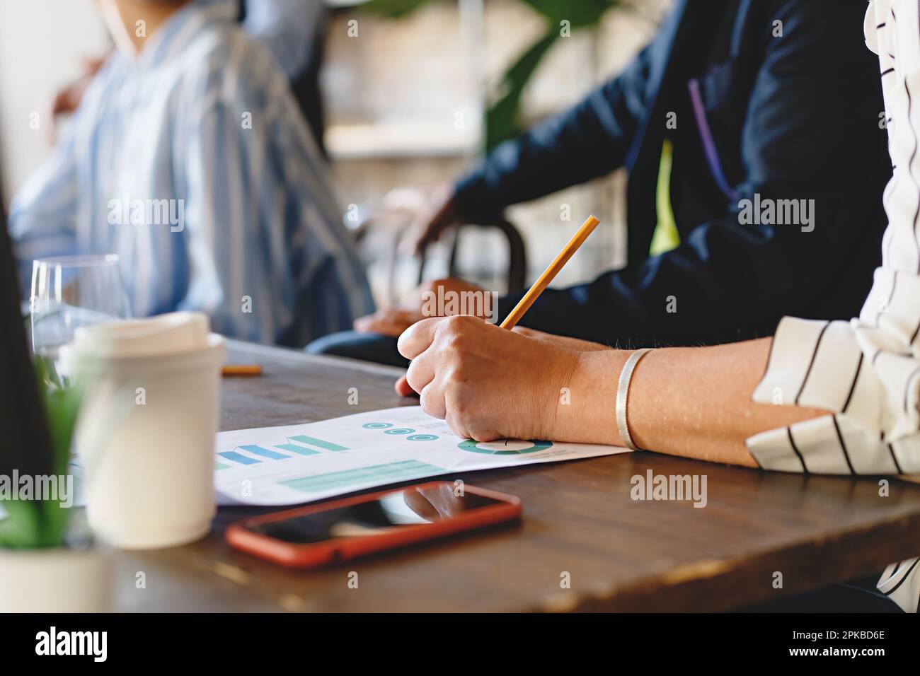 Primo piano della mano di una donna che prende appunti su carta da tavolo durante la riunione in ufficio, tavolo di legno con altre persone, documenti, smartphone e caffè. Foto Stock