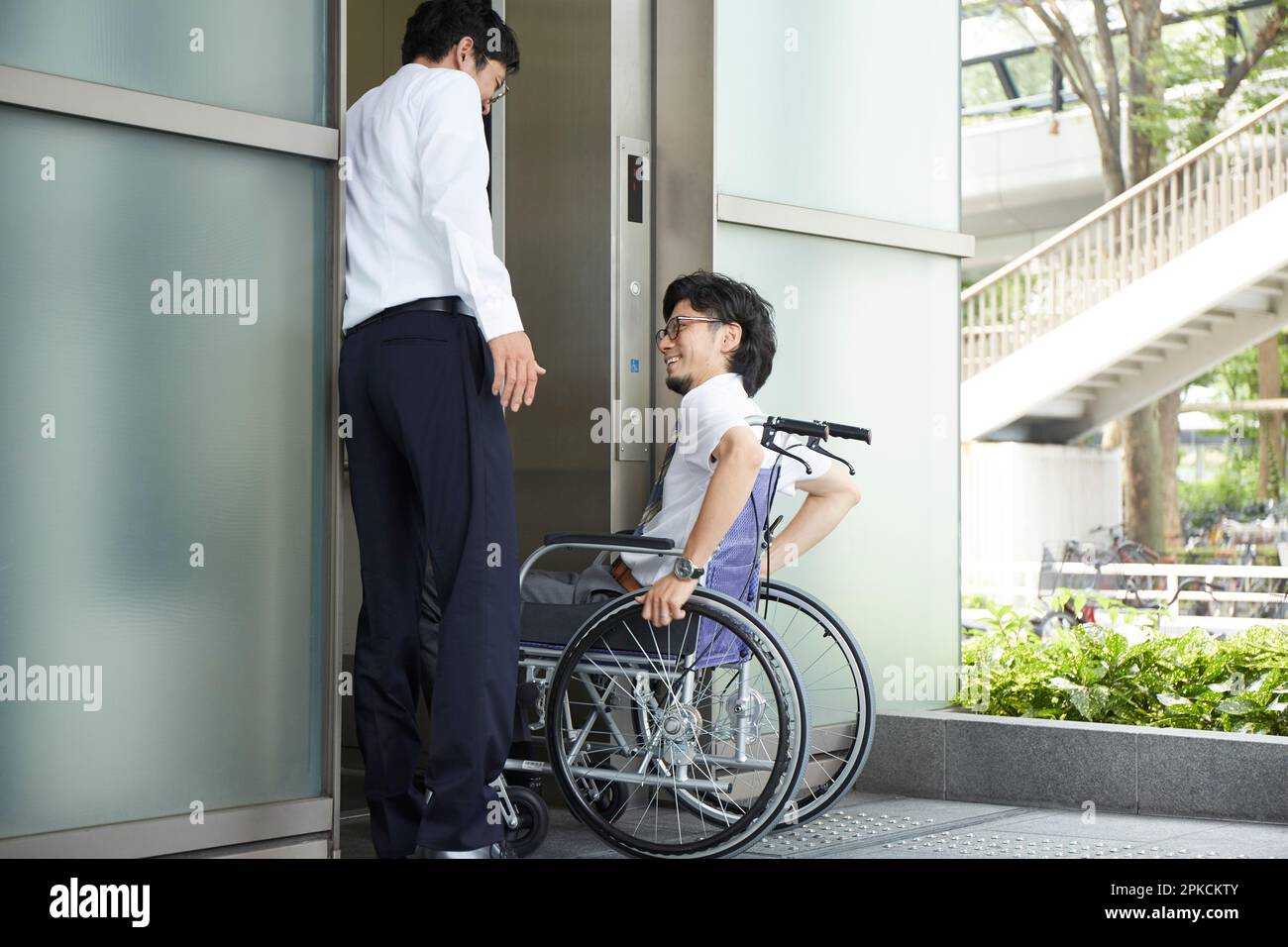 Un uomo in sedia a rotelle che cerca di salire sull'ascensore e un uomo che lo aiuta Foto Stock