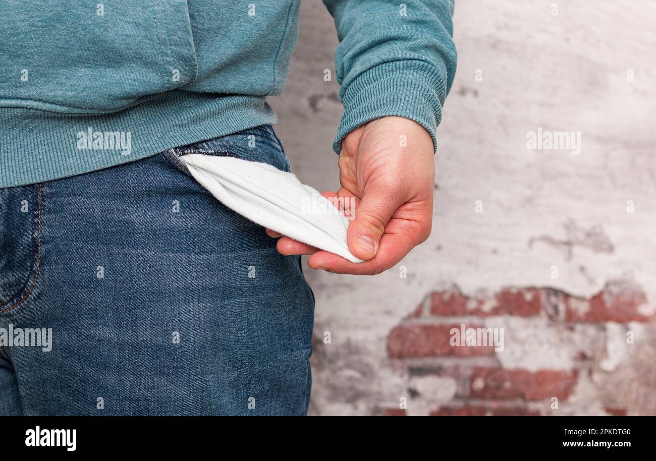 Primo piano di una mano di un uomo caucasico irriconoscibile che indossa jeans che mostra la tasca vuota dei suoi pantaloni davanti a una vecchia parete. Foto Stock