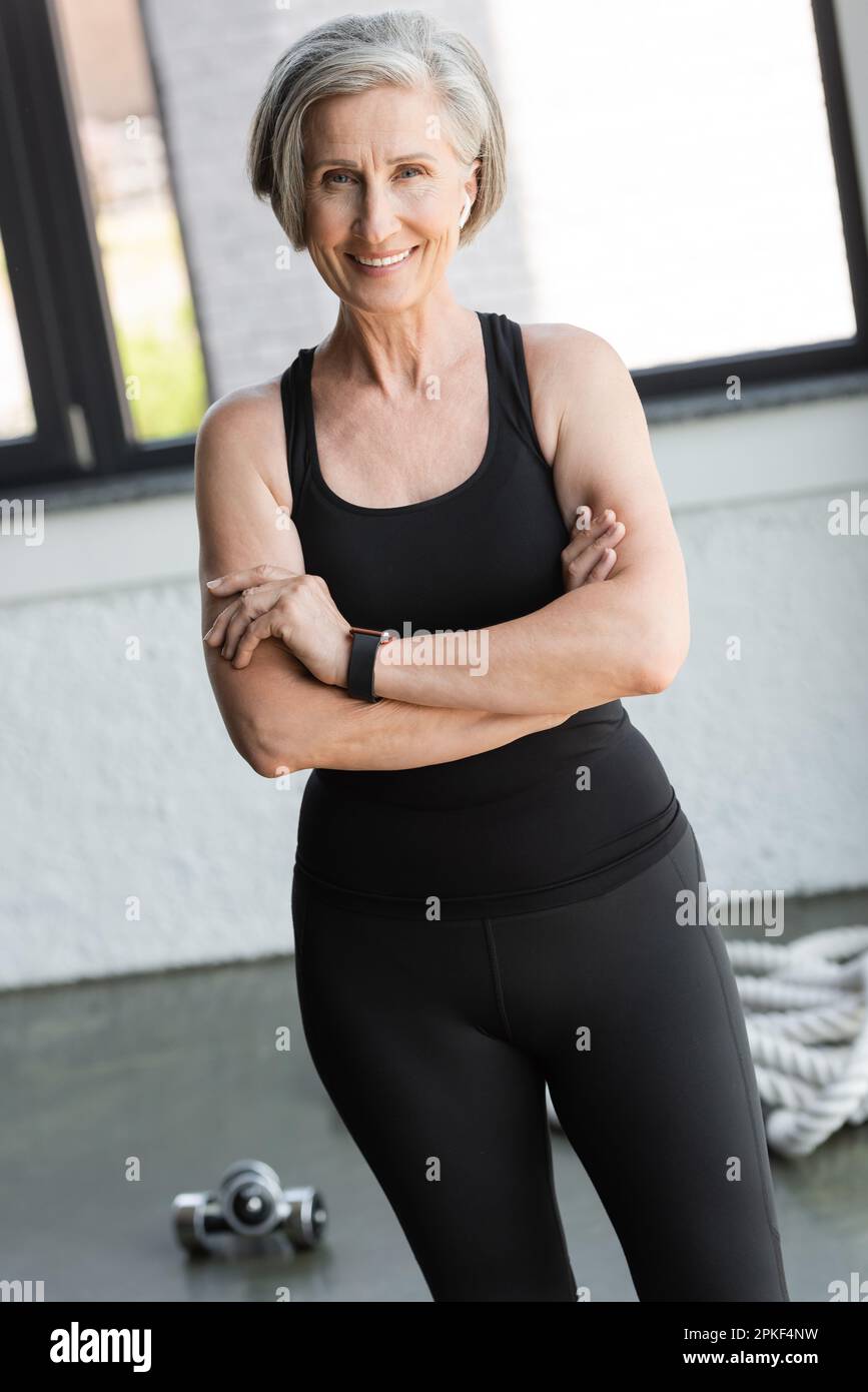 donna in forma e pensionato in canotta sorridente mentre in piedi con braccia incrociate vicino attrezzature sportive in palestra, immagine stock Foto Stock