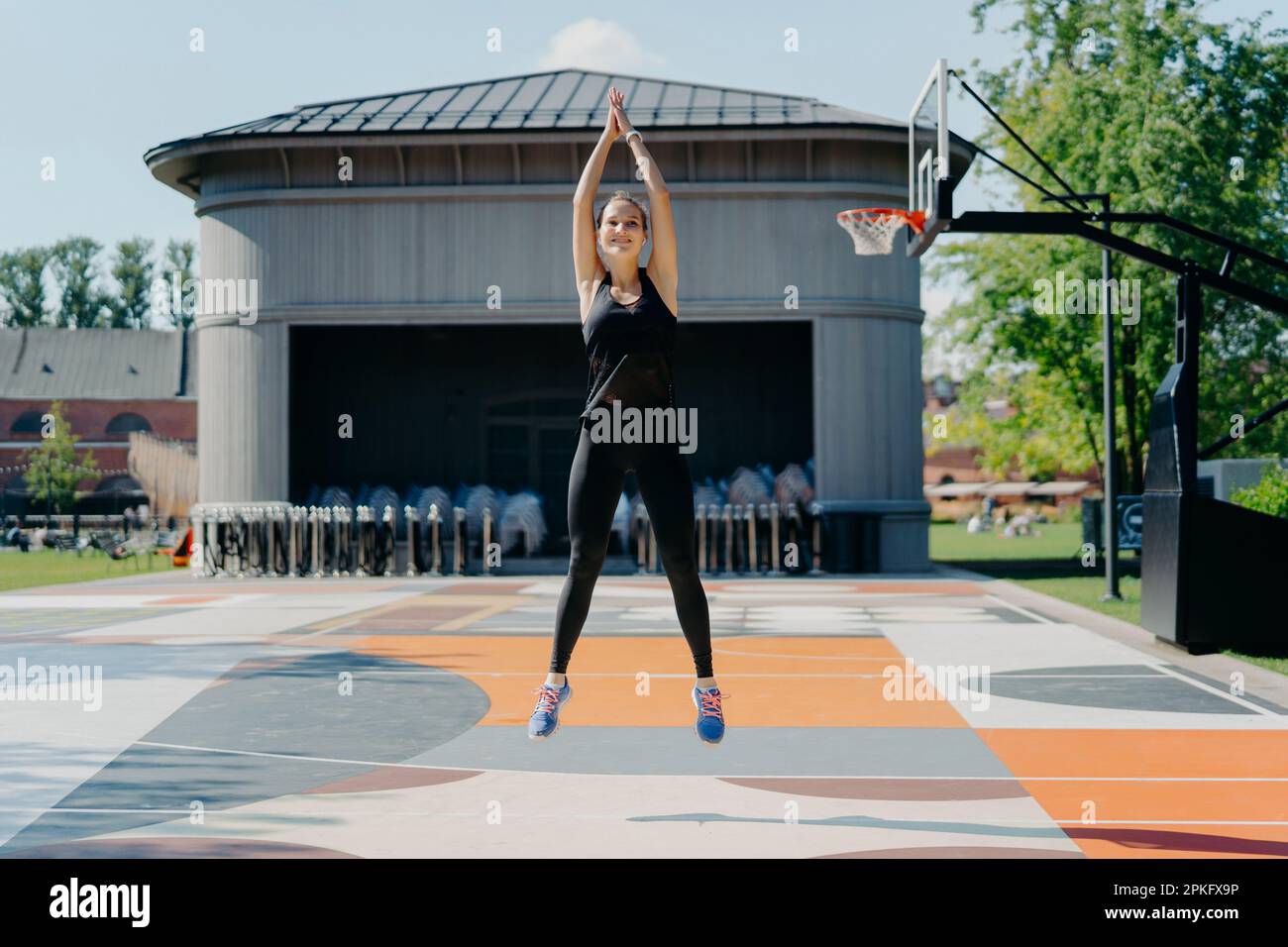 Atletica giovane donna in nero Activewear salti in alto mantiene le braccia sollevate sui ganci le mani si scalda prima di jogging ha allenamento allo stadio all'aperto ha auto Foto Stock
