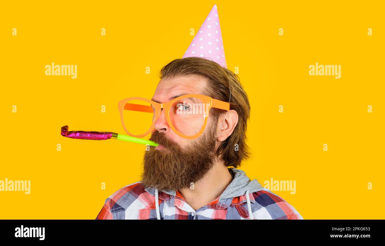 Feste e celebrazioni. Uomo bearded in cappello di compleanno e divertenti occhiali che soffiano il fischio di festa. Buon compleanno. Bell'uomo in cappello conico e grande Foto Stock