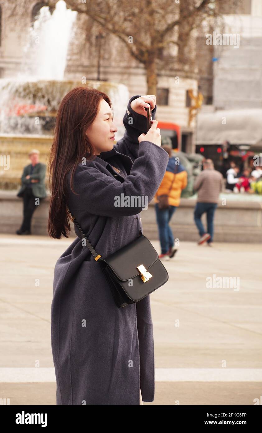 Una giovane donna dell'Estremo Oriente che prende un selfie a Trafalgar Square, Londra. Regno Unito con una borsa nera e un lungo cappotto grigio Foto Stock