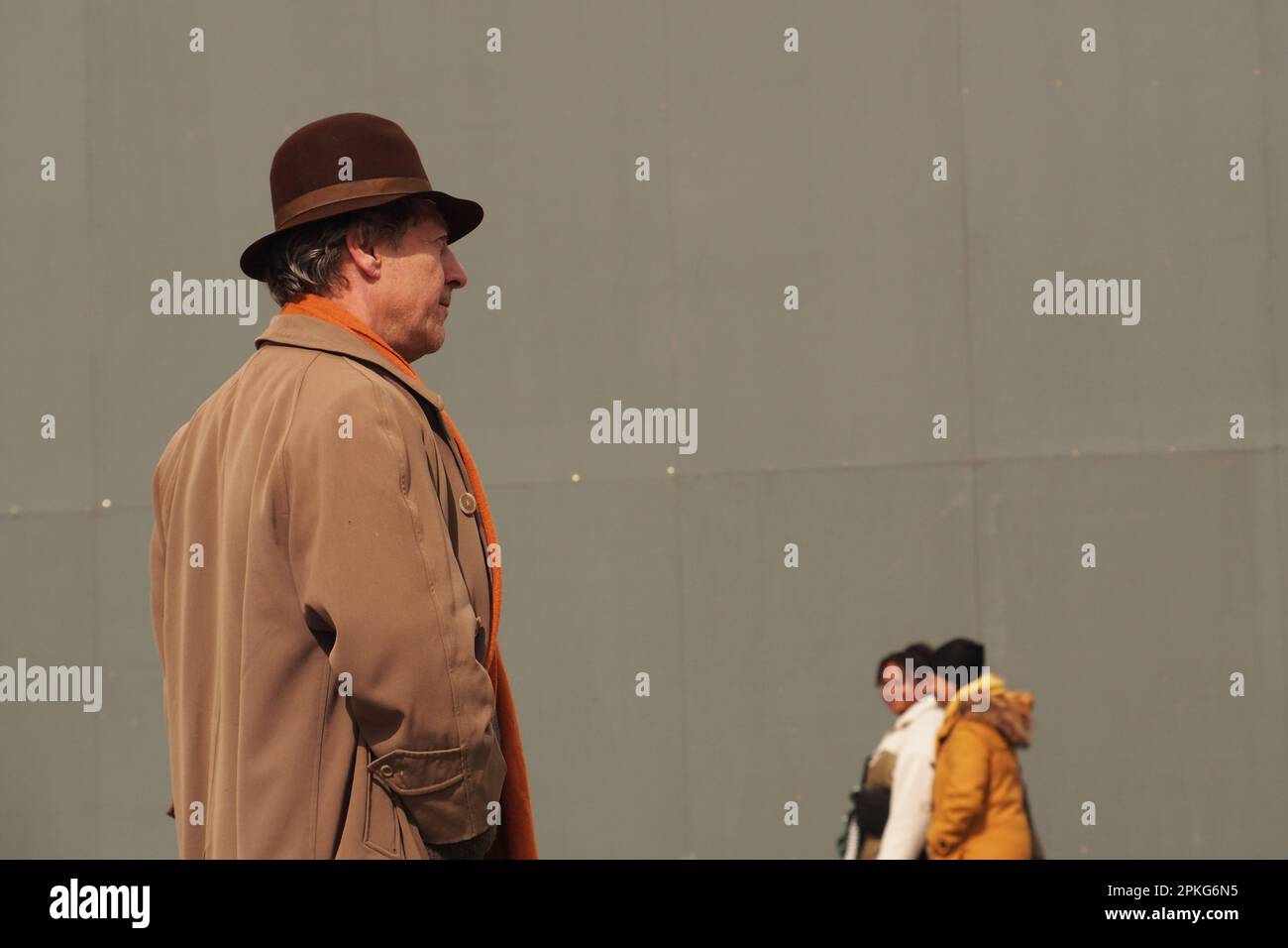 Vista laterale di un uomo 60+, dalla vita in alto, in Trafalgar Square, Londra. Regno Unito indossa un cappello marrone, un soprabito marrone e una sciarpa arancione, guardando Foto Stock