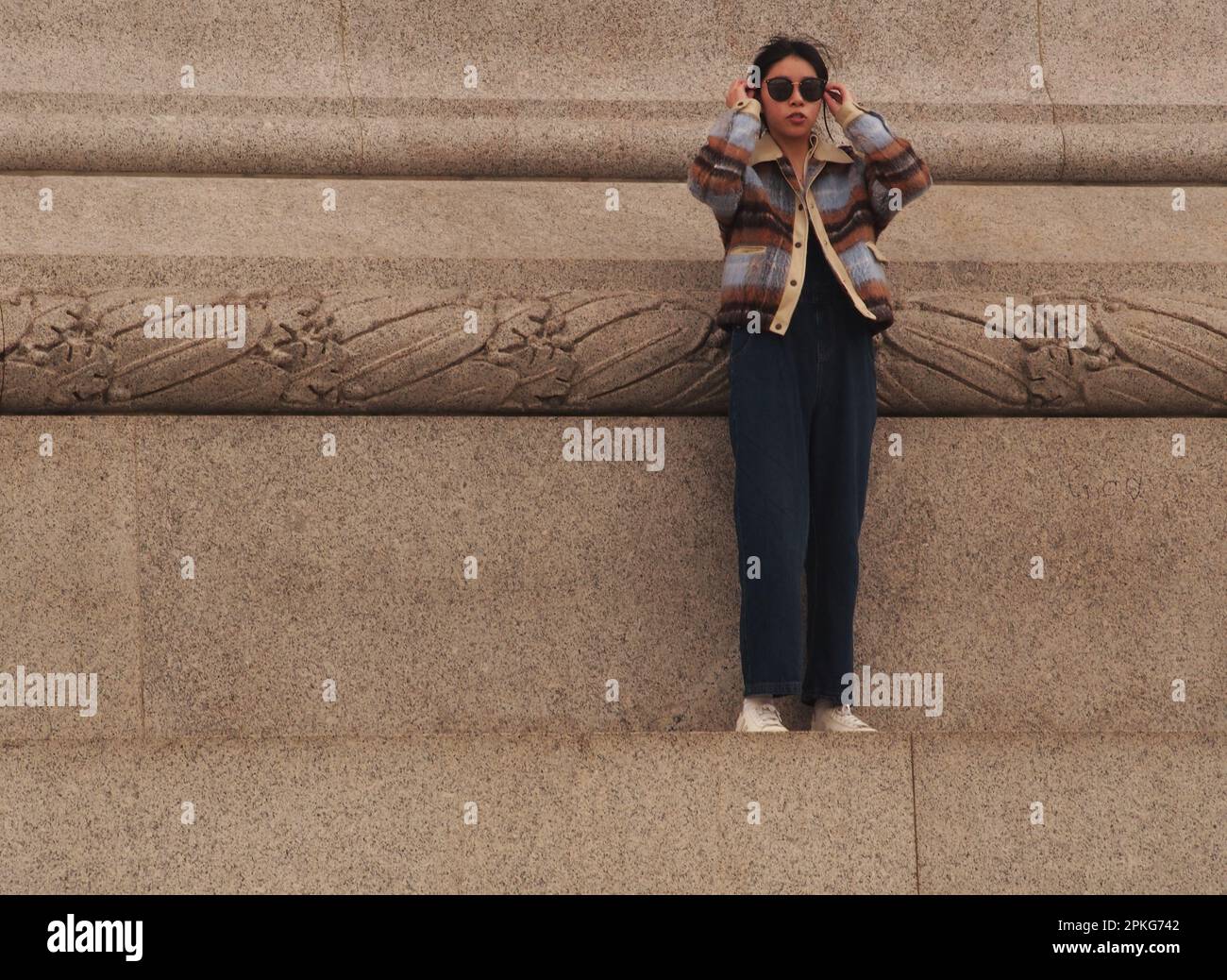 Una giovane donna dell'Asia orientale che si erge da sola sul plinto che circonda la colonna di Nelson a Trafalgar Square, Londra, Inghilterra. REGNO UNITO Foto Stock