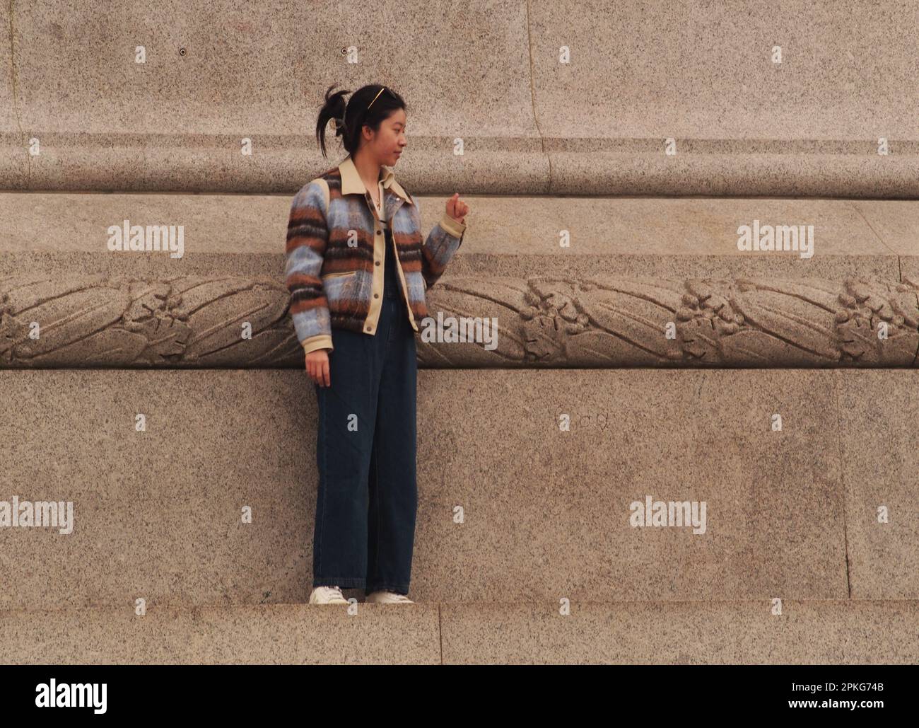Una giovane donna dell'Asia orientale che si erge da sola sul plinto che circonda la colonna di Nelson a Trafalgar Square, Londra, Inghilterra. REGNO UNITO Foto Stock