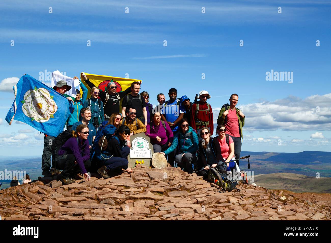 UK Weather: Venerdì Santo, 7th aprile 2023. Brecon Beacons National Park, Galles del Sud. Un gruppo di escursionisti di Manchester che detengono le bandiere del Lancashire e dello Yorkshire hanno fatto scattare la loro foto sul punto di trig sulla cima della Pen y Fan nel Parco Nazionale Brecon Beacons. Pen y Fan (886m / 2.907ft) è la vetta più alta del Brecon Beacons National Park e nel sud della Gran Bretagna e una popolare destinazione escursionistica. Il bel tempo soleggiato ha fatto sì che molte persone si sono fatte fare il viaggio al Parco per le vacanze di banca di oggi. Foto Stock