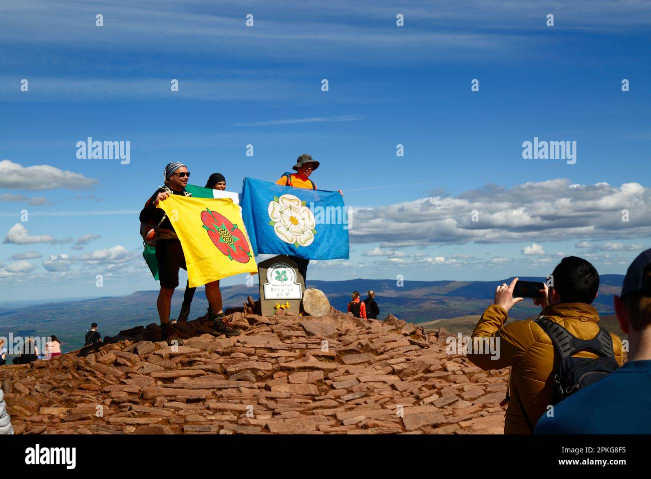 UK Weather: Venerdì Santo, 7th aprile 2023. Brecon Beacons National Park, Galles del Sud. Gli escursionisti che detengono le bandiere del Lancashire e dello Yorkshire hanno la loro foto scattata sul punto di trig sulla cima di Pen y Fan nel Brecon Beacons National Park. Pen y Fan (886m / 2.907ft) è la vetta più alta del Brecon Beacons National Park e nel sud della Gran Bretagna e una popolare destinazione escursionistica. Il bel tempo soleggiato ha fatto sì che molte persone si sono fatte fare il viaggio al Parco per le vacanze di banca di oggi. Foto Stock