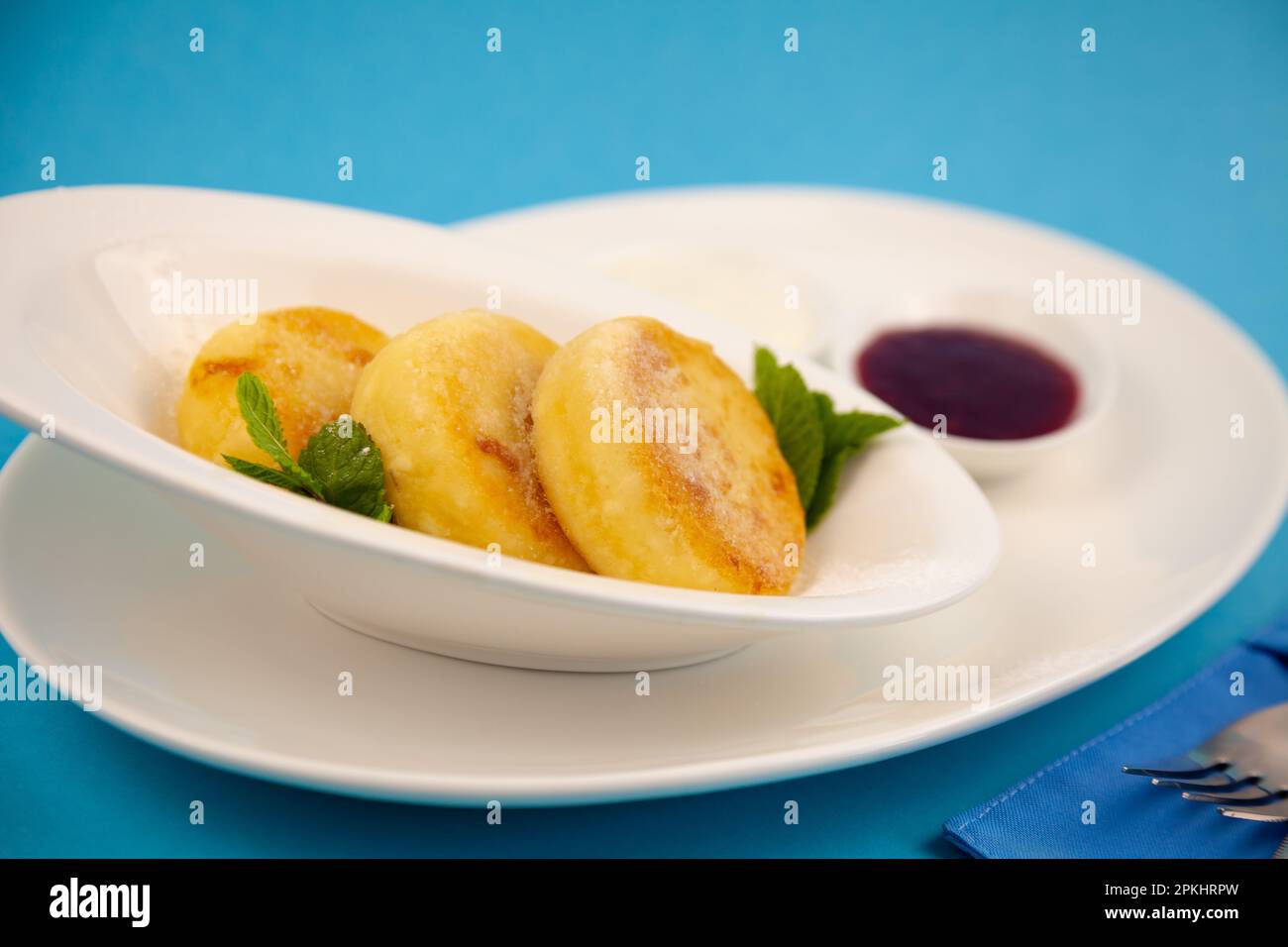 Frittelle di formaggio ricotta rotonda in un piatto bianco su sfondo blu. Cibo prima colazione. Cibo sano. Salsa di panna acida e menta. Primo piano vista dall'alto. Pasto dessert. Frittelle di ricotta al formaggio ricotta. Foto Stock